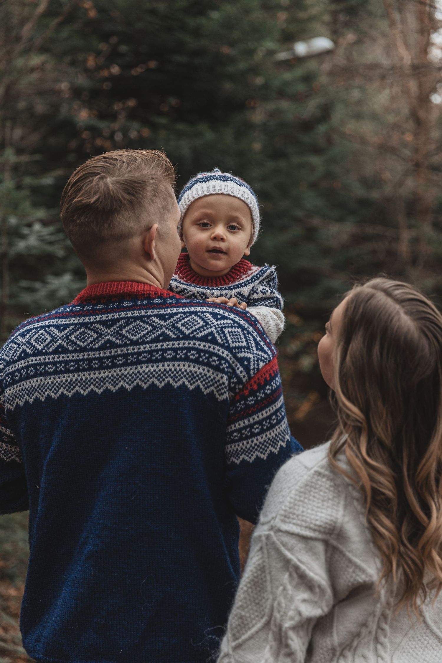 Family wearing cozy winter sweaters with Nordic patterns stands together in a forest setting during autumn.
