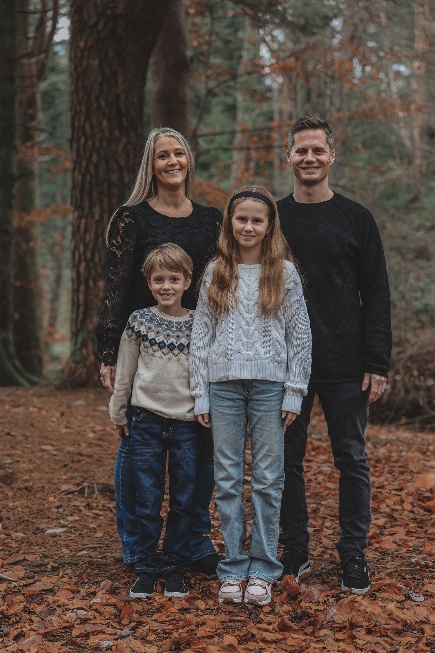 A family of four wearing cozy sweaters poses together on fallen leaves in an autumn forest setting.