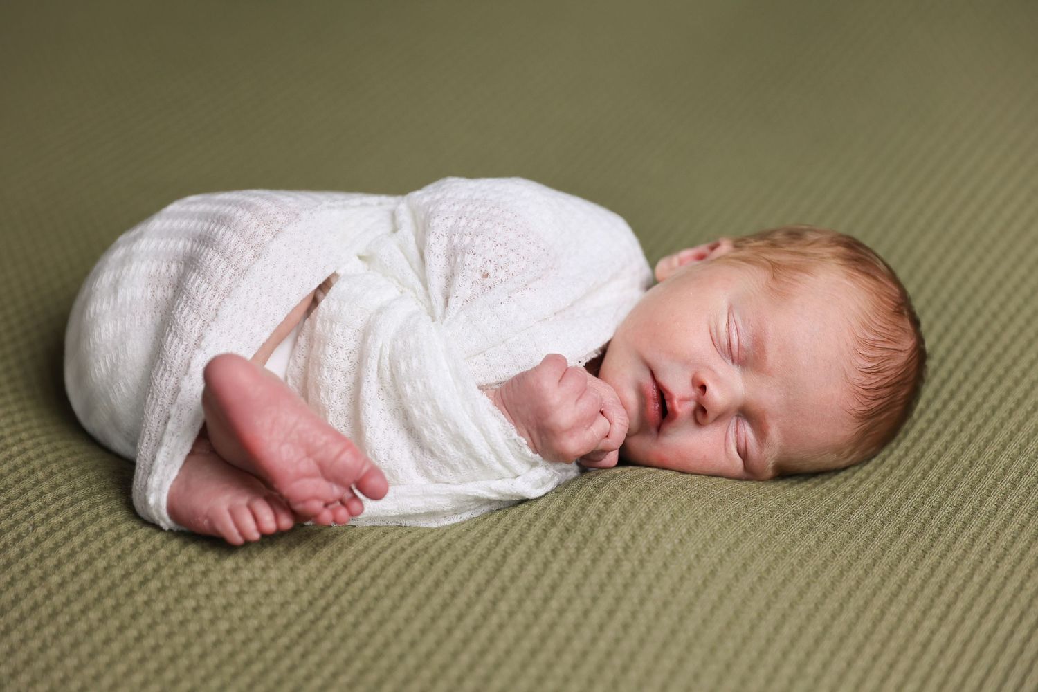 Sleeping newborn wrapped in white knit blanket on sage green background.