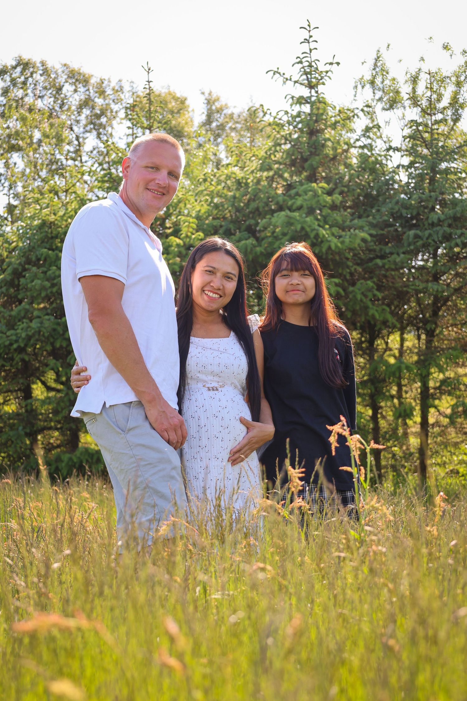 Three people stand together in a sunlit meadow with tall grass and trees in the background.