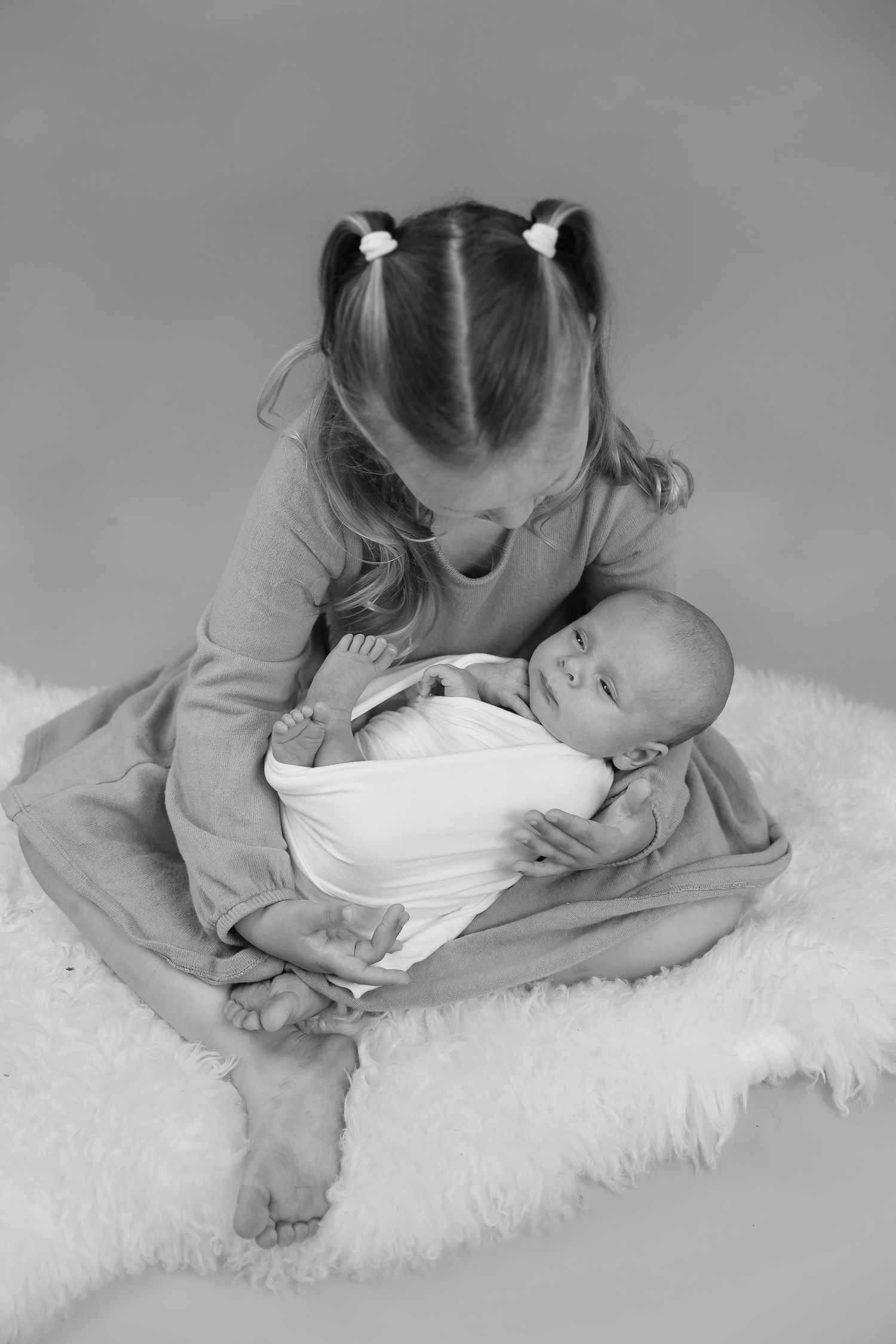 A young child in pigtails gently holds and gazes down at a newborn baby on a white fluffy rug.