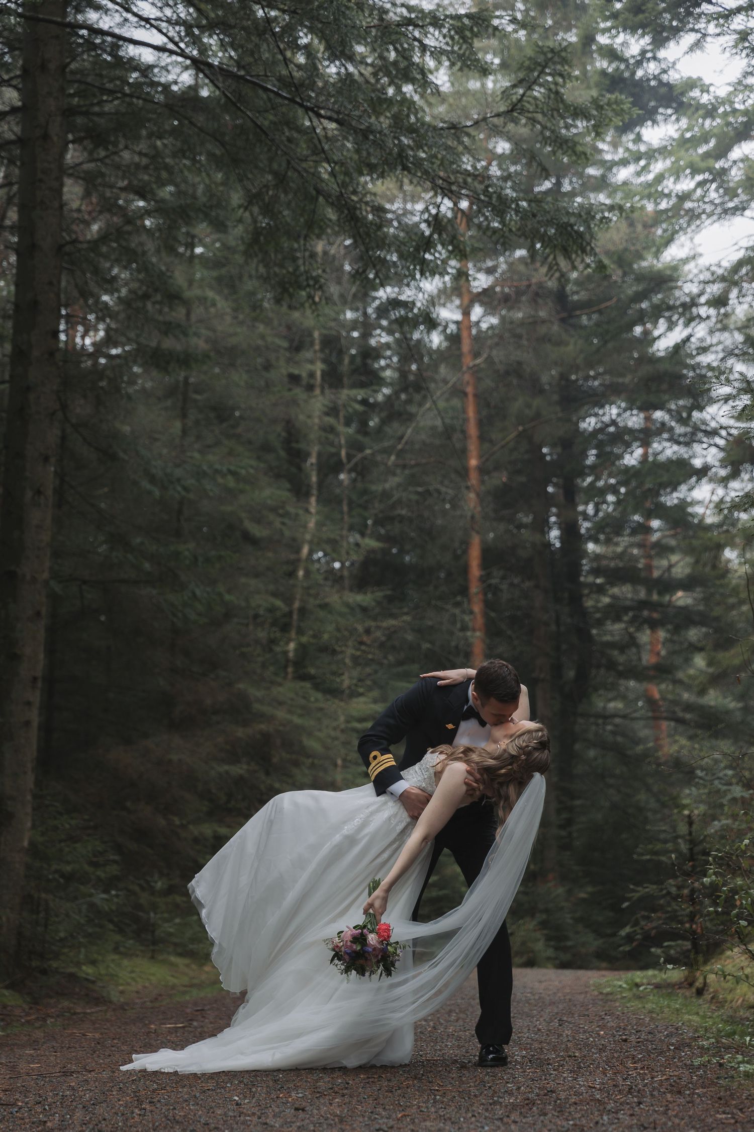 Bride and groom share a romantic dip kiss on a forest path, with the wedding dress flowing gracefully.
