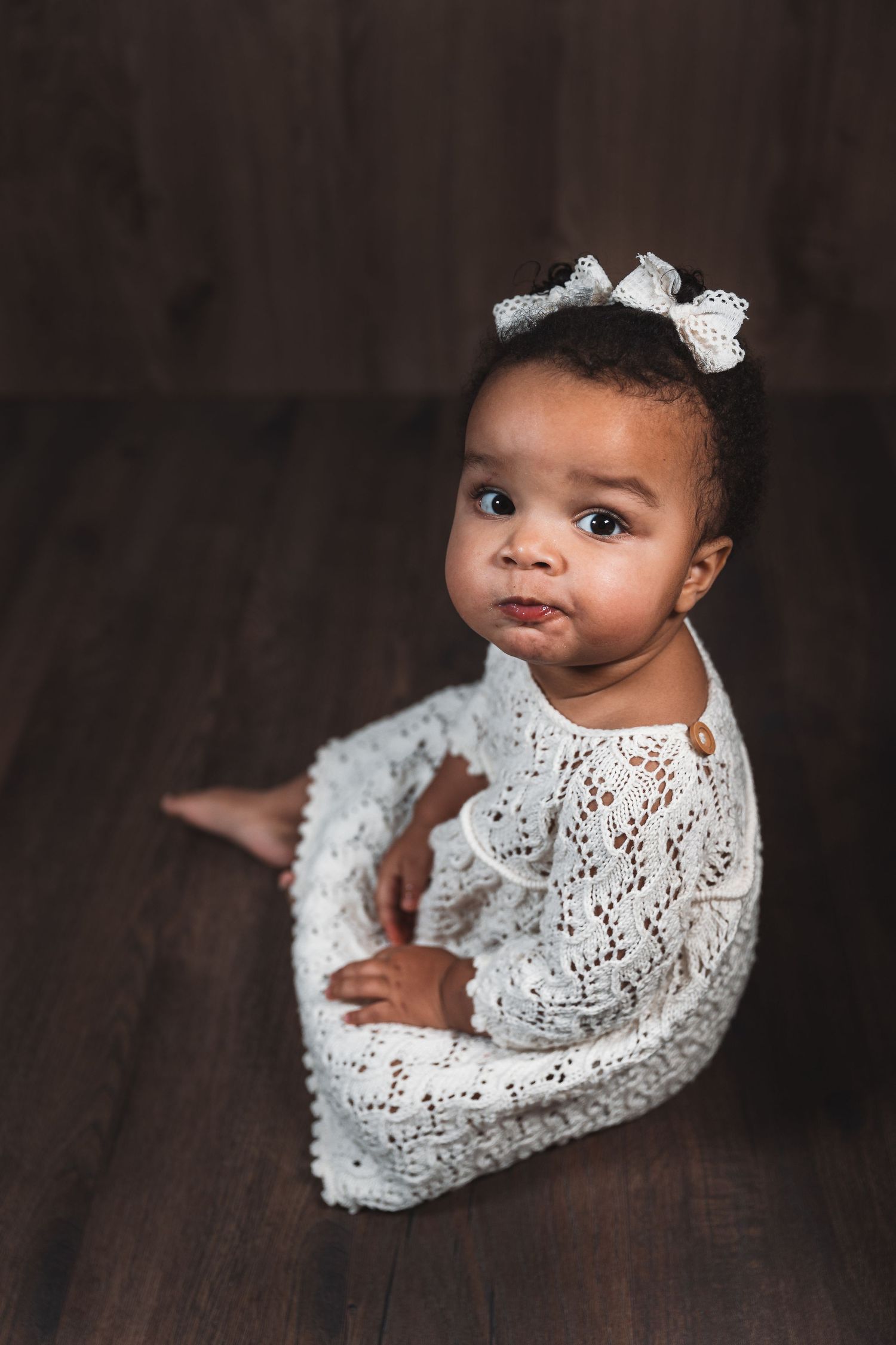 Baby wearing white lace dress and bow sitting on wooden floor.