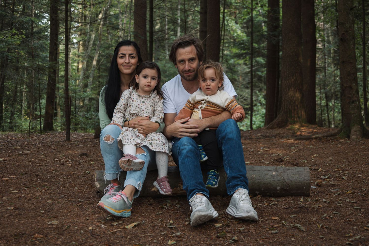 A family of four sits together on a log in a peaceful forest setting.