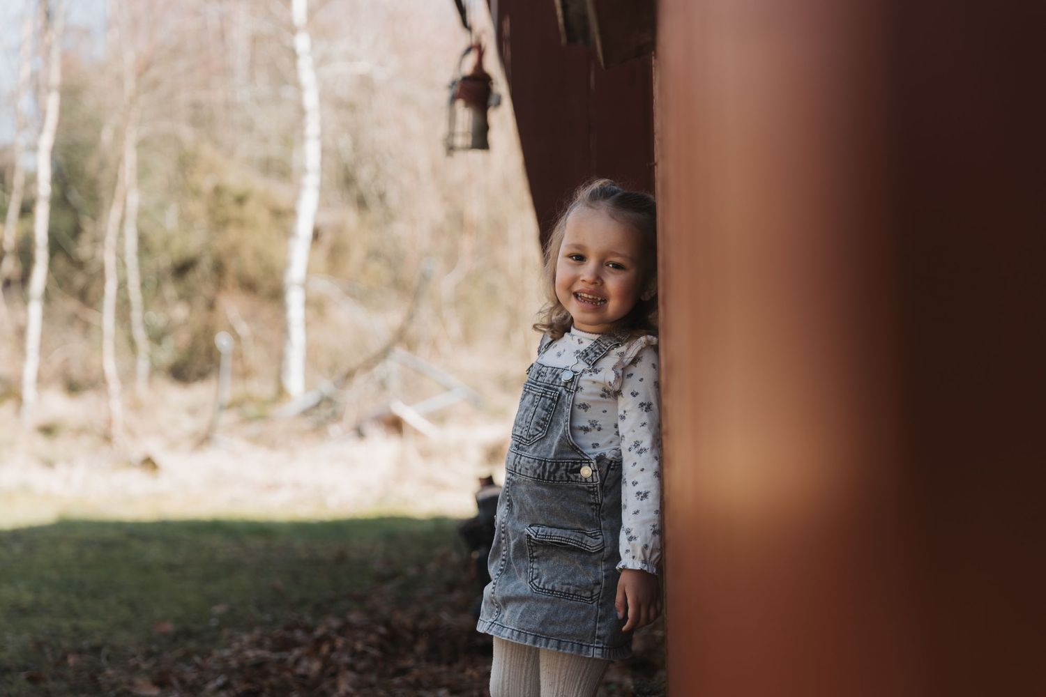 A small child wearing a gray dress and floral top peeks around a wooden pillar in an outdoor autumn setting.