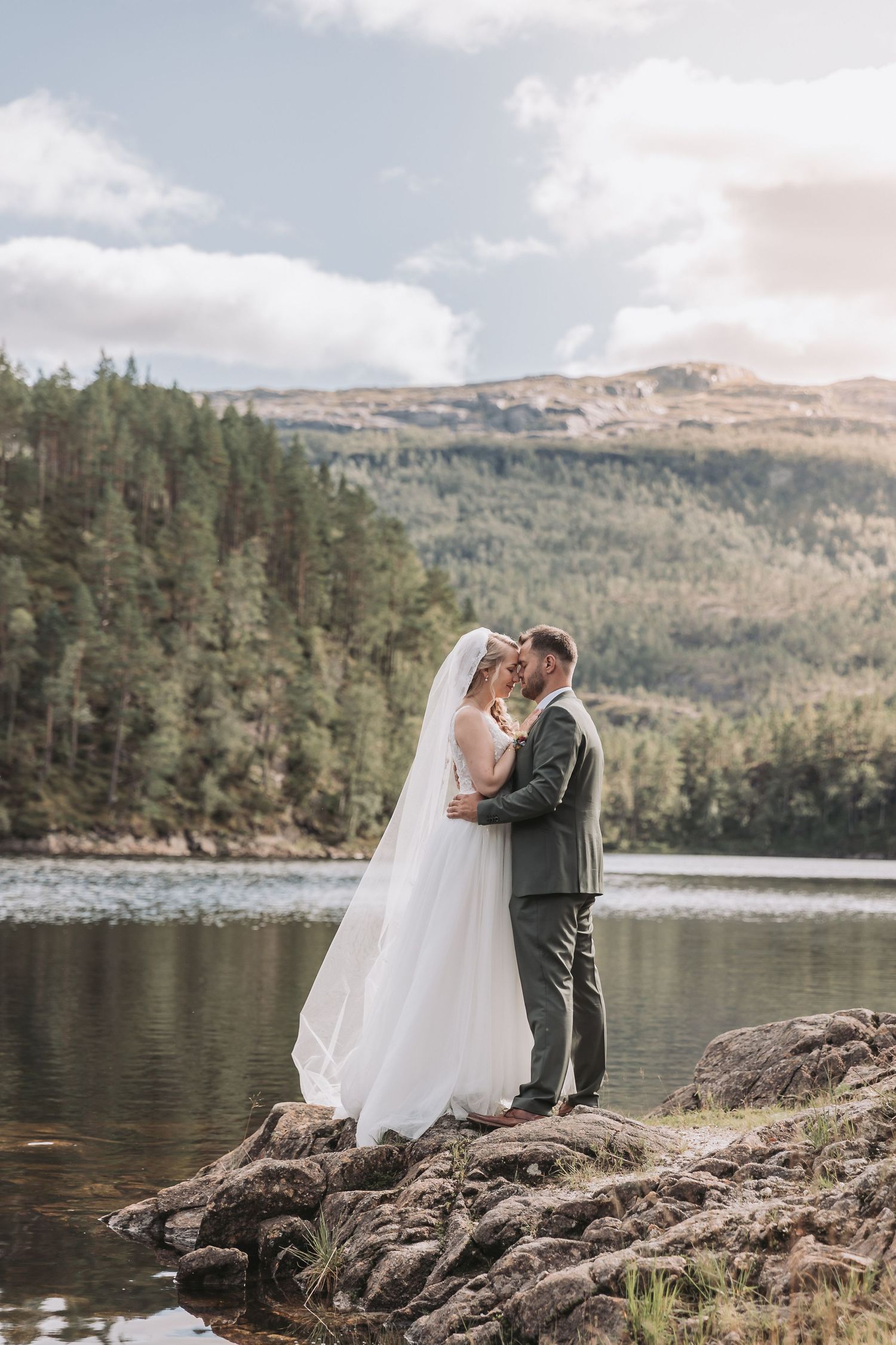 Bride and groom embrace on rocky lakeside with forested mountains backdrop on their wedding day.