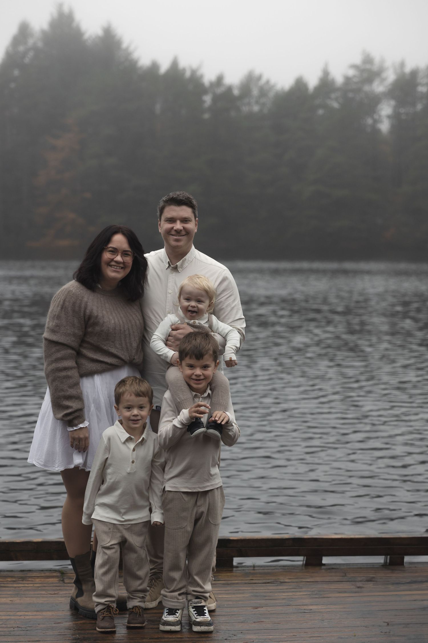 Family of five posing on a misty lakeside dock with tall evergreen trees in the background.
