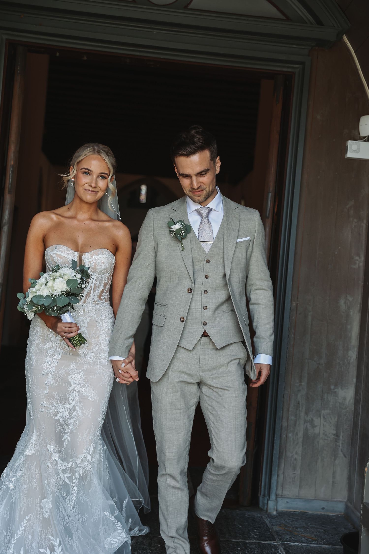 Bride in lace mermaid gown holding bouquet walks with groom in grey suit exiting church doors on wedding day.