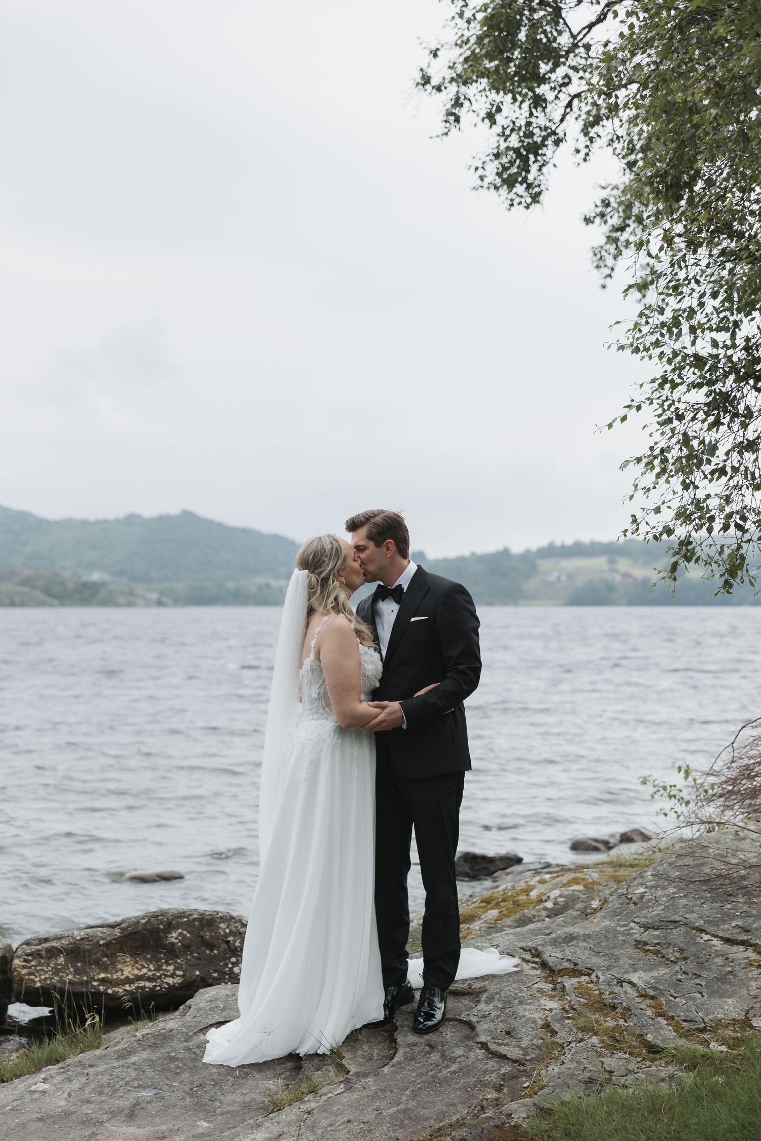 Bride and groom share a romantic kiss by a lakeside, dressed in wedding attire with misty mountains in the background.