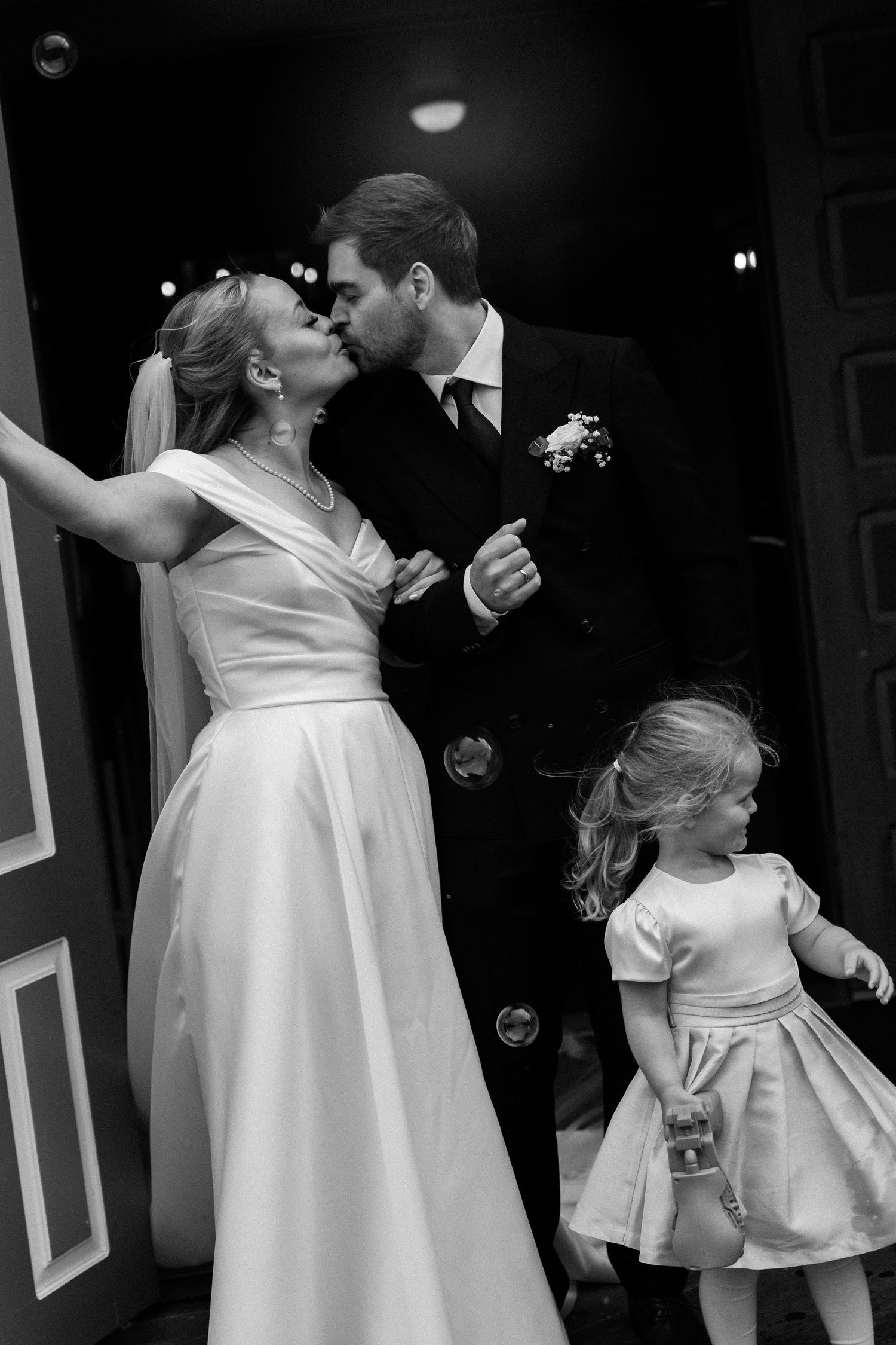 Black and white photo of a bride and groom kissing at their wedding while a young flower girl plays nearby.