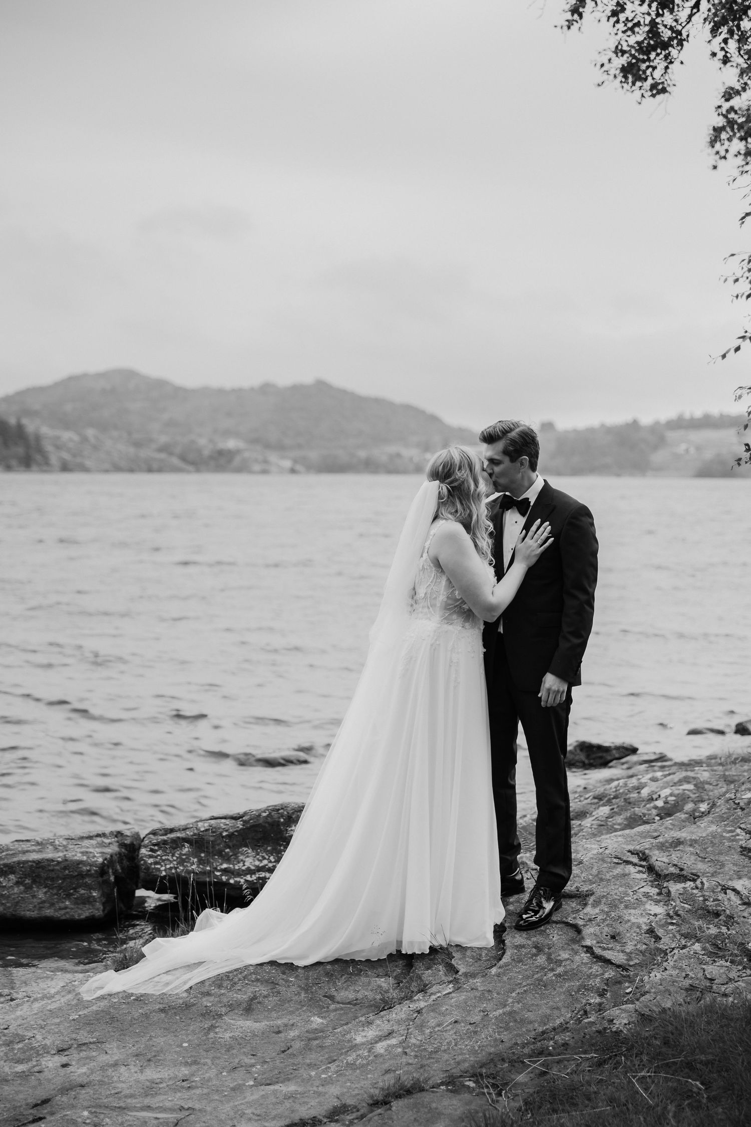 Black and white photo of a bride and groom sharing a romantic moment by a scenic lake with mountain views.