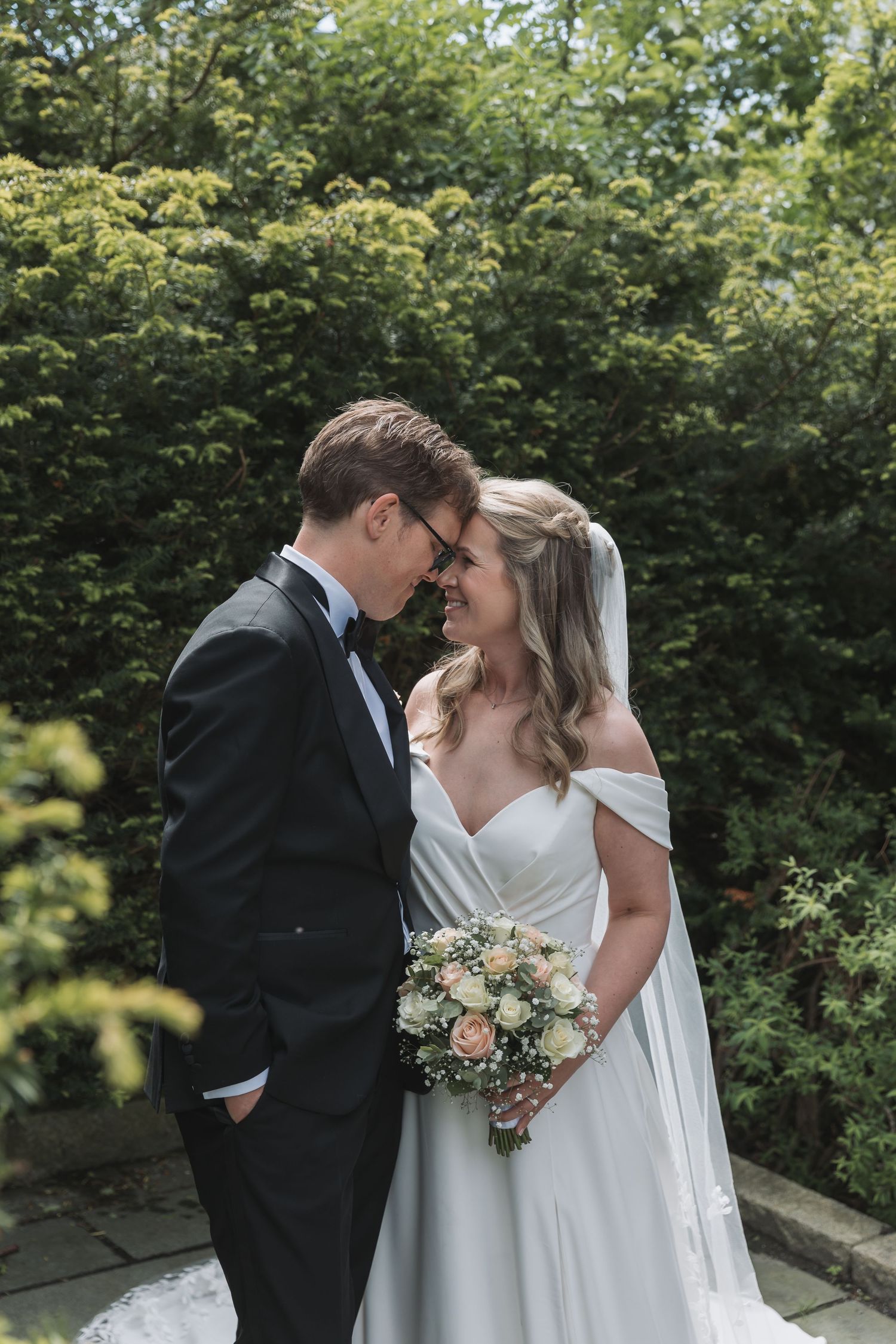 Smiling bride and groom touching foreheads in a lush garden, bride holding a bouquet.