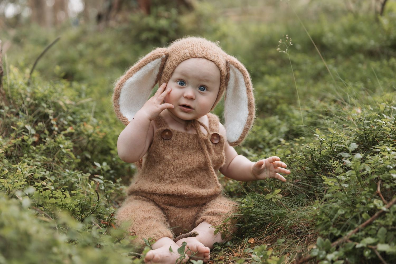 A baby wearing a brown bunny costume with long floppy ears sits in green grass during an outdoor photoshoot.