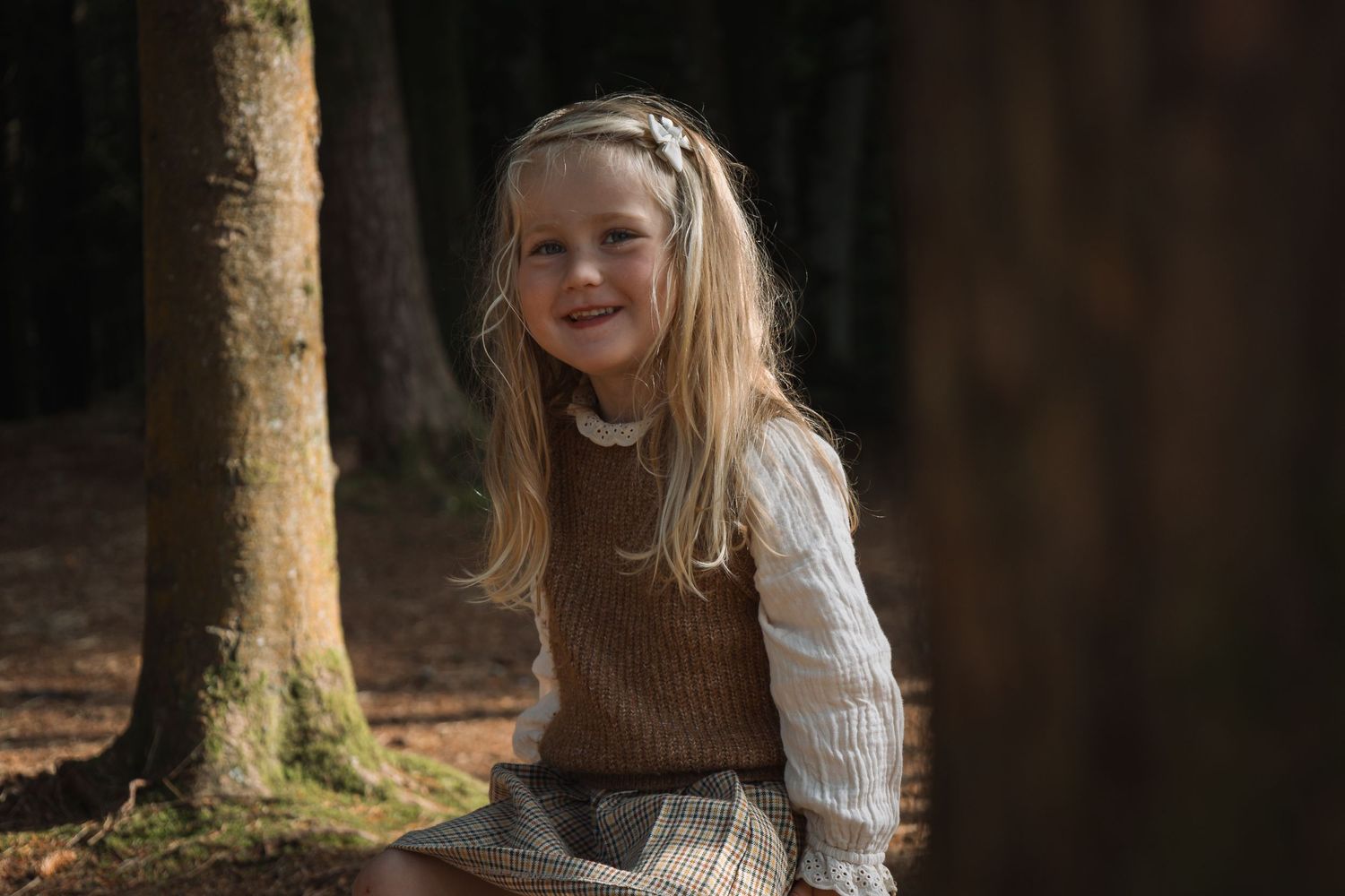 Child in brown and cream sweater sits among trees in dappled forest light.