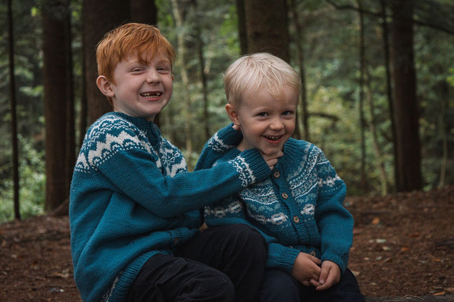 Two children wearing matching blue patterned sweaters share a joyful laugh in the woods.