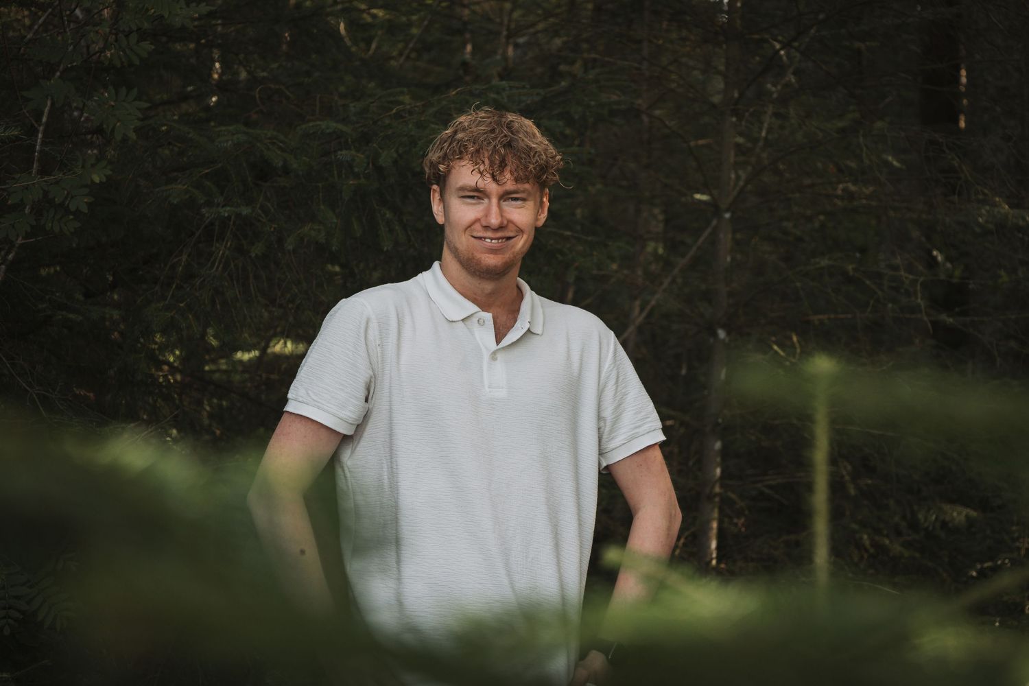 Portrait in white polo shirt standing outdoors against dark forest background.