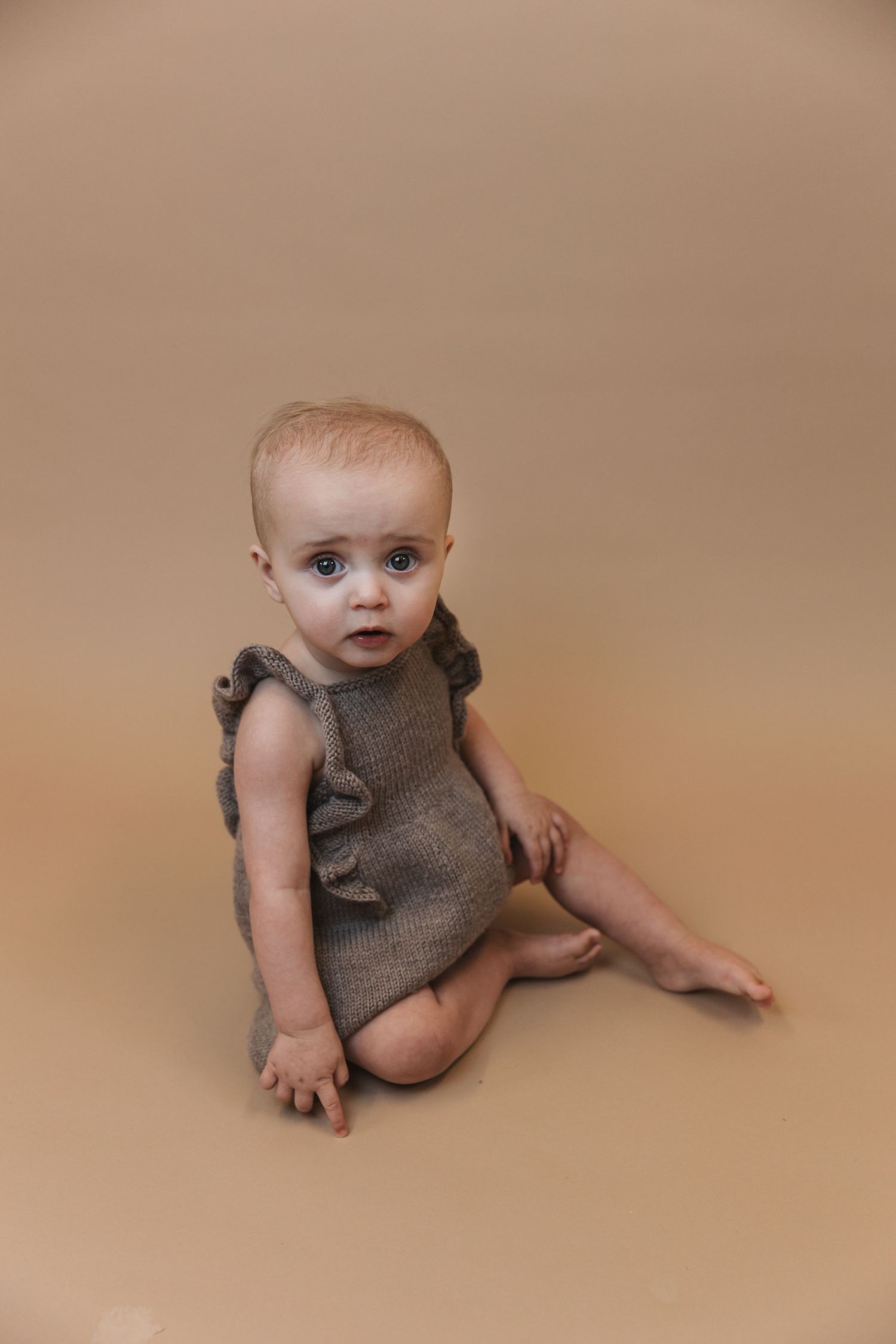 Baby wearing a gray sleeveless romper sits on beige background during professional photo session.