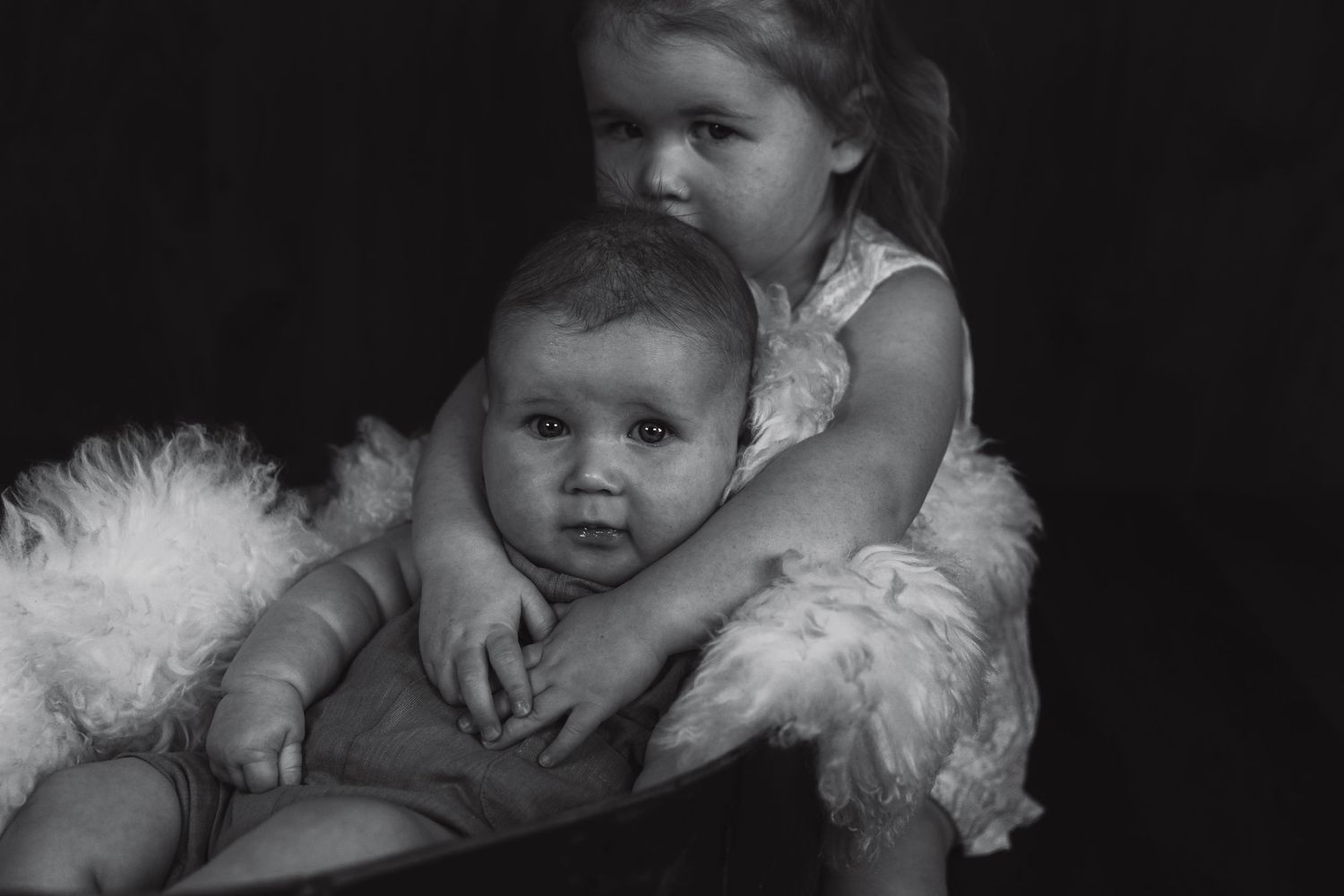 A black and white portrait of two young children in white dresses posing together against a dark background.