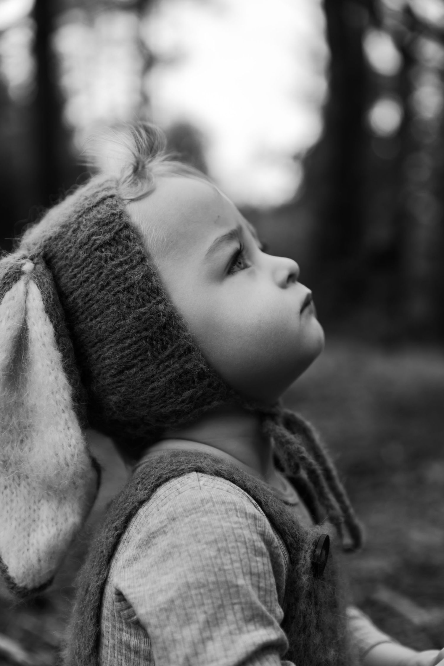 Black and white portrait of a young child wearing a knit bunny ear hat, gazing upward in a forest setting.