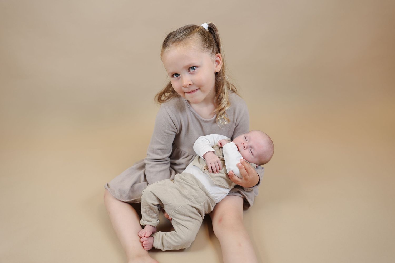 A young child in a beige outfit sits on a neutral backdrop holding a small baby wearing light-colored clothing.