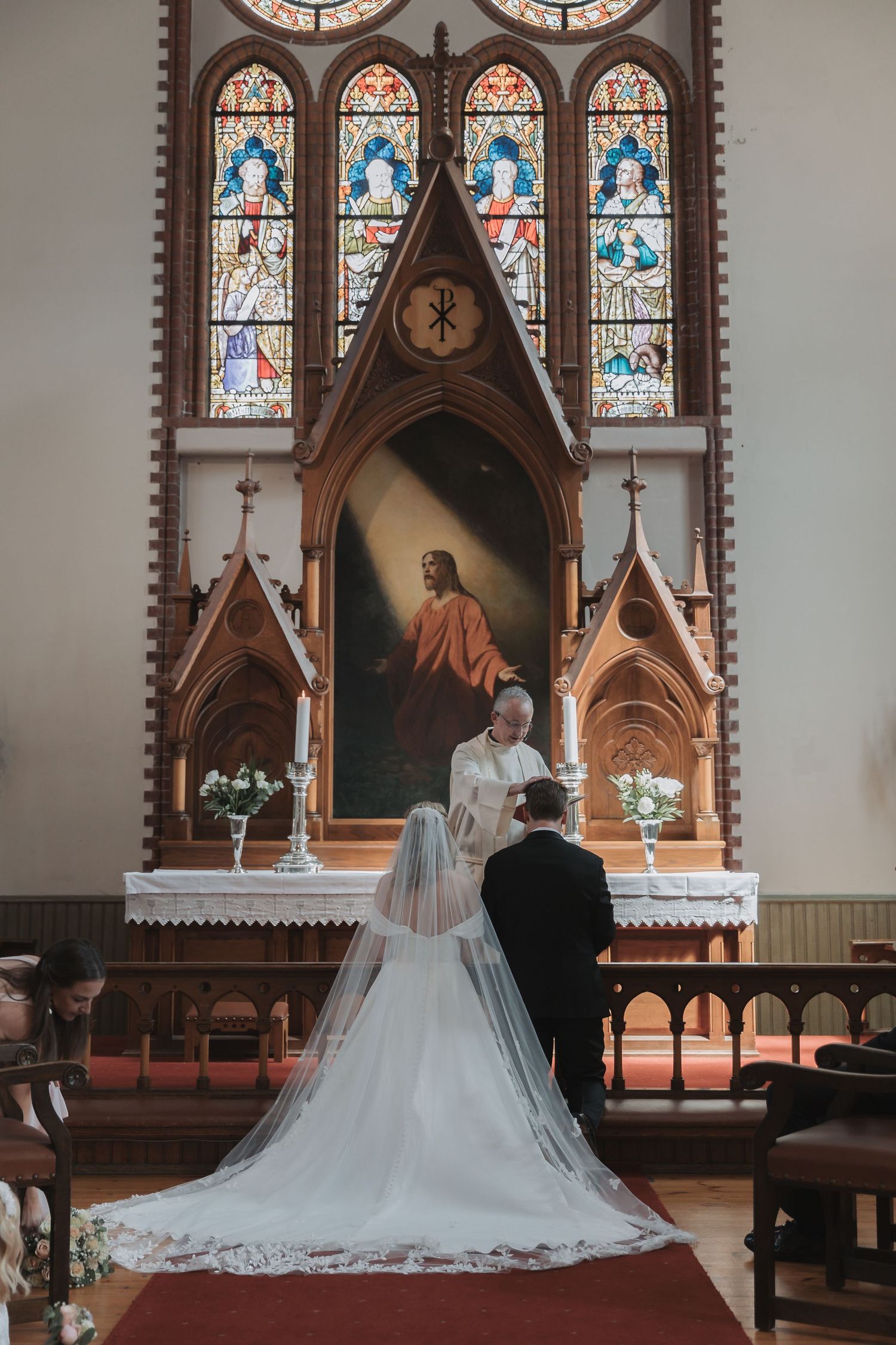 Bride and groom exchange vows at a church altar with stained glass windows and a painting of Jesus.