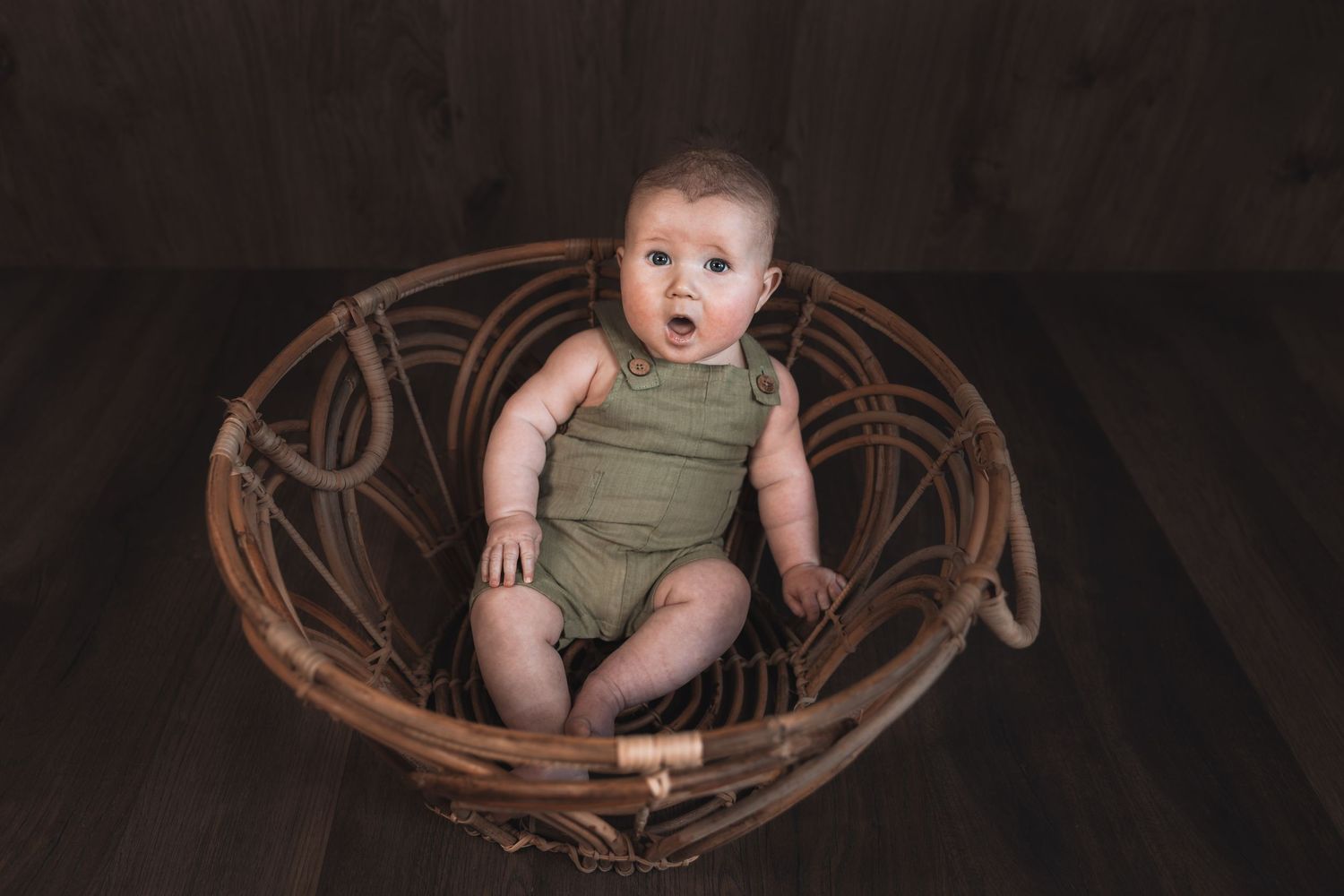 Baby wearing olive green romper sits in round woven bamboo basket against dark backdrop.
