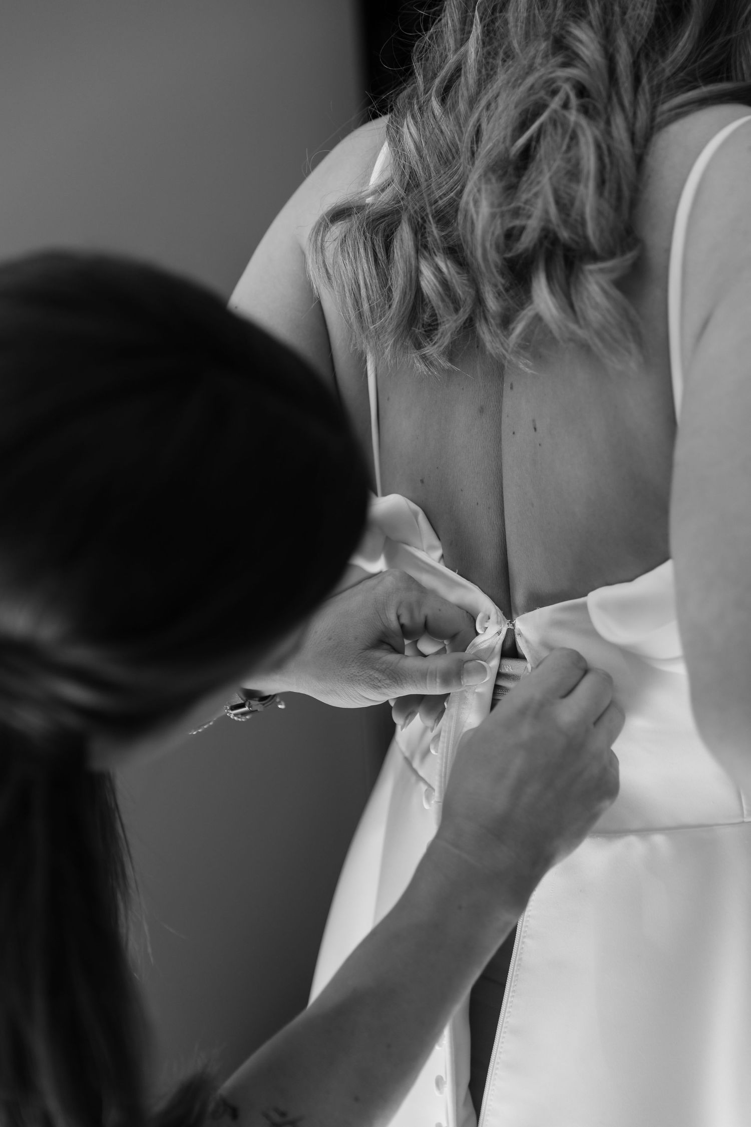 Black and white photo of a bridesmaid buttoning the back of a bride's wedding dress before the ceremony.