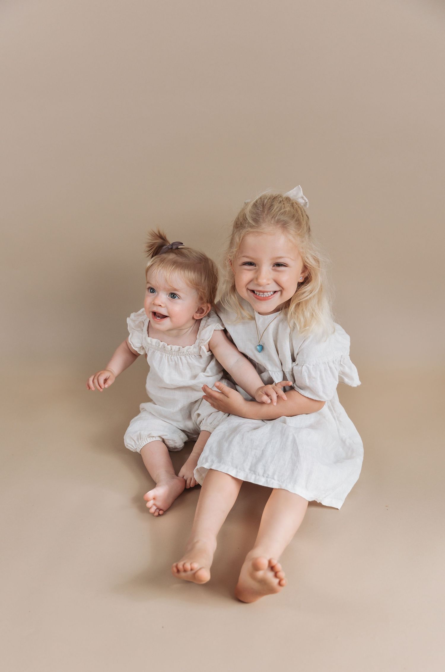 Two young sisters in white dresses sit together on a beige backdrop, the older girl smiling and hugging the baby.
