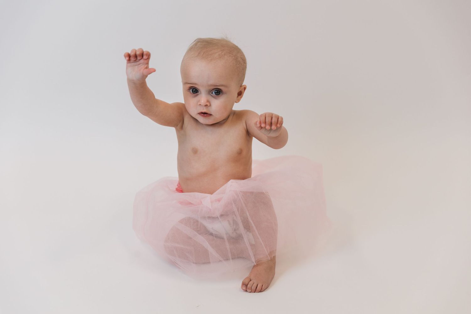 Small baby in pink tulle skirt sits against white background with arms raised in playful pose.