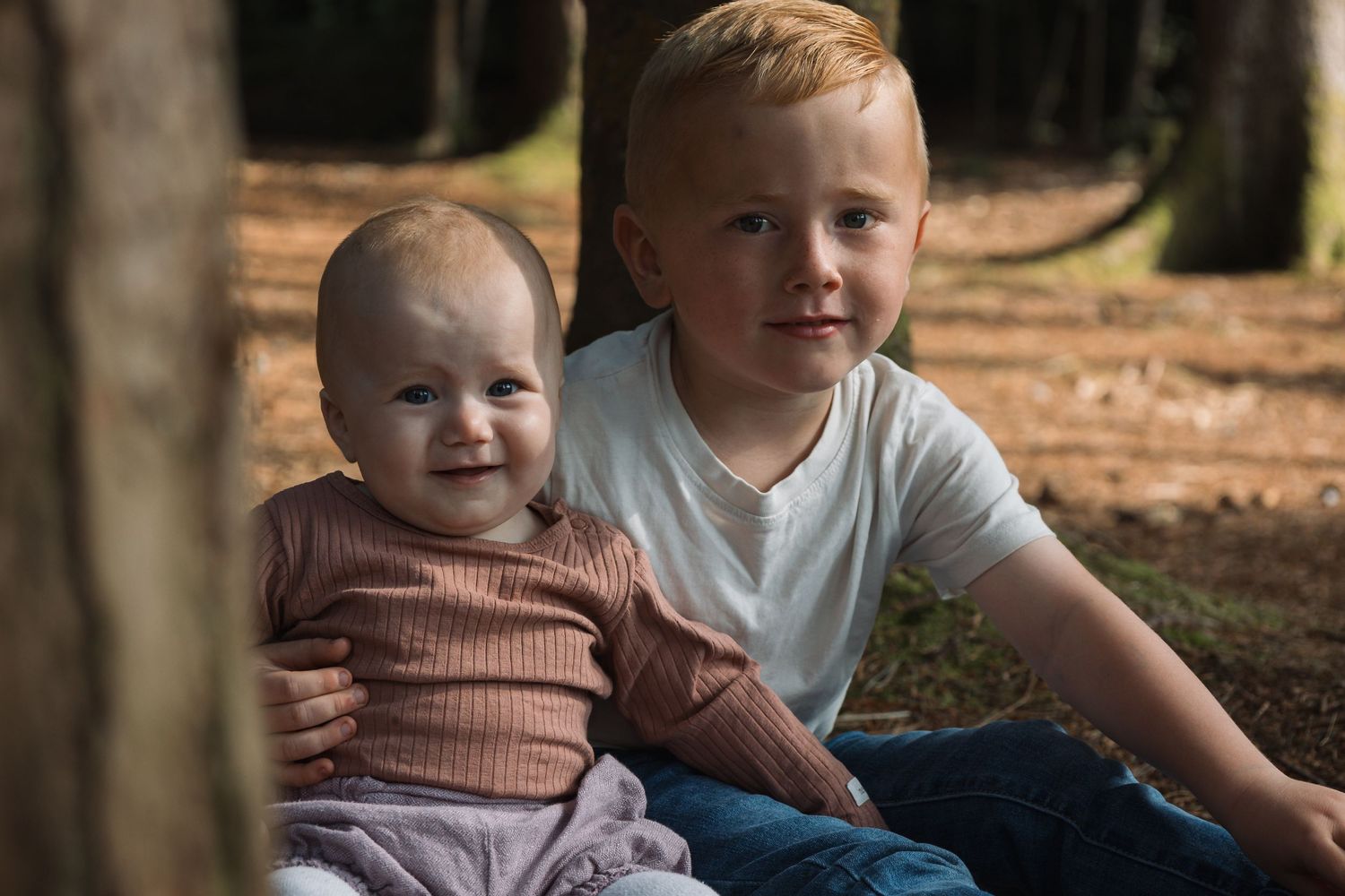 Two siblings sitting together outdoors against a wooded backdrop in casual attire.