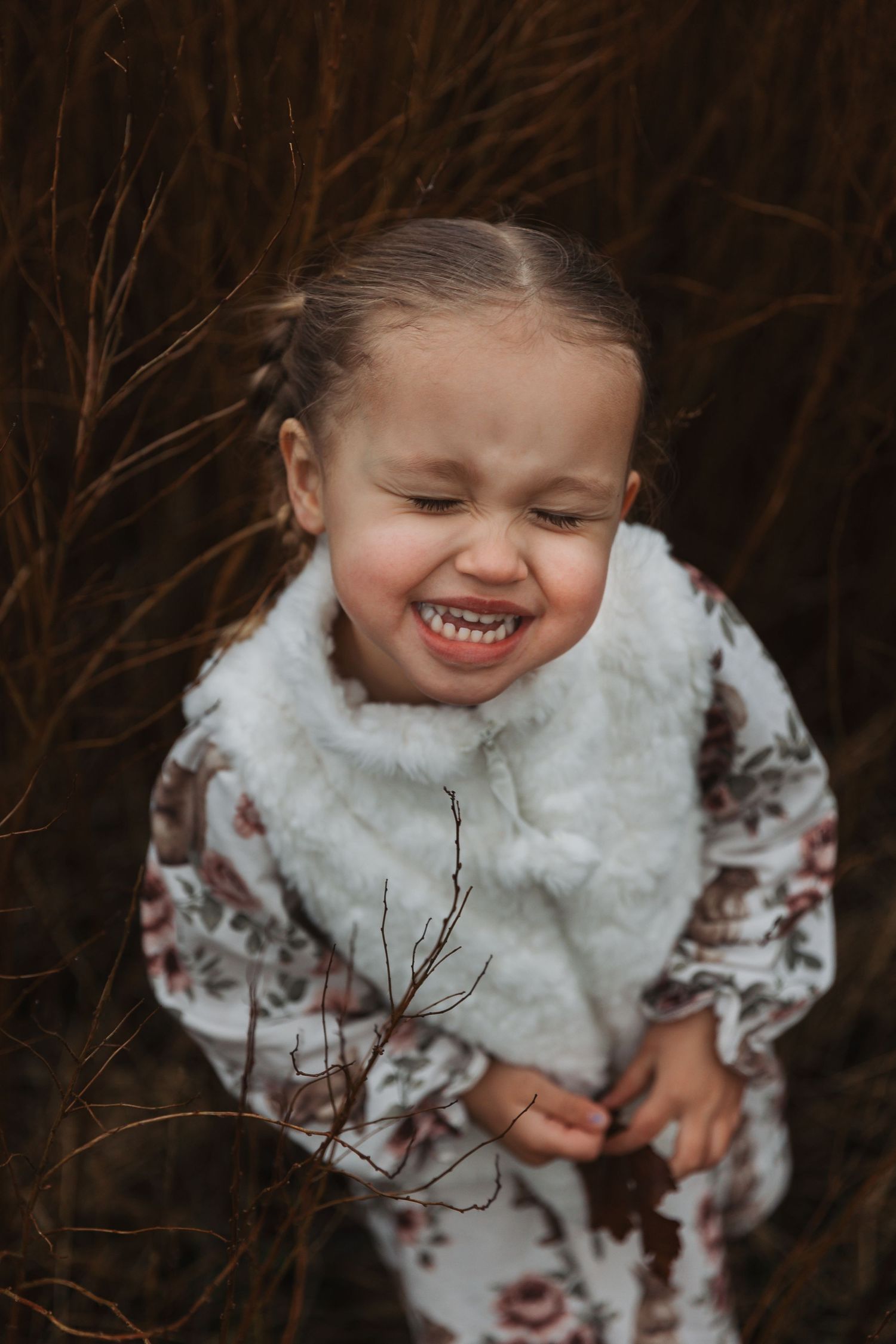 Child in white fur vest laughing joyfully against dark natural background.