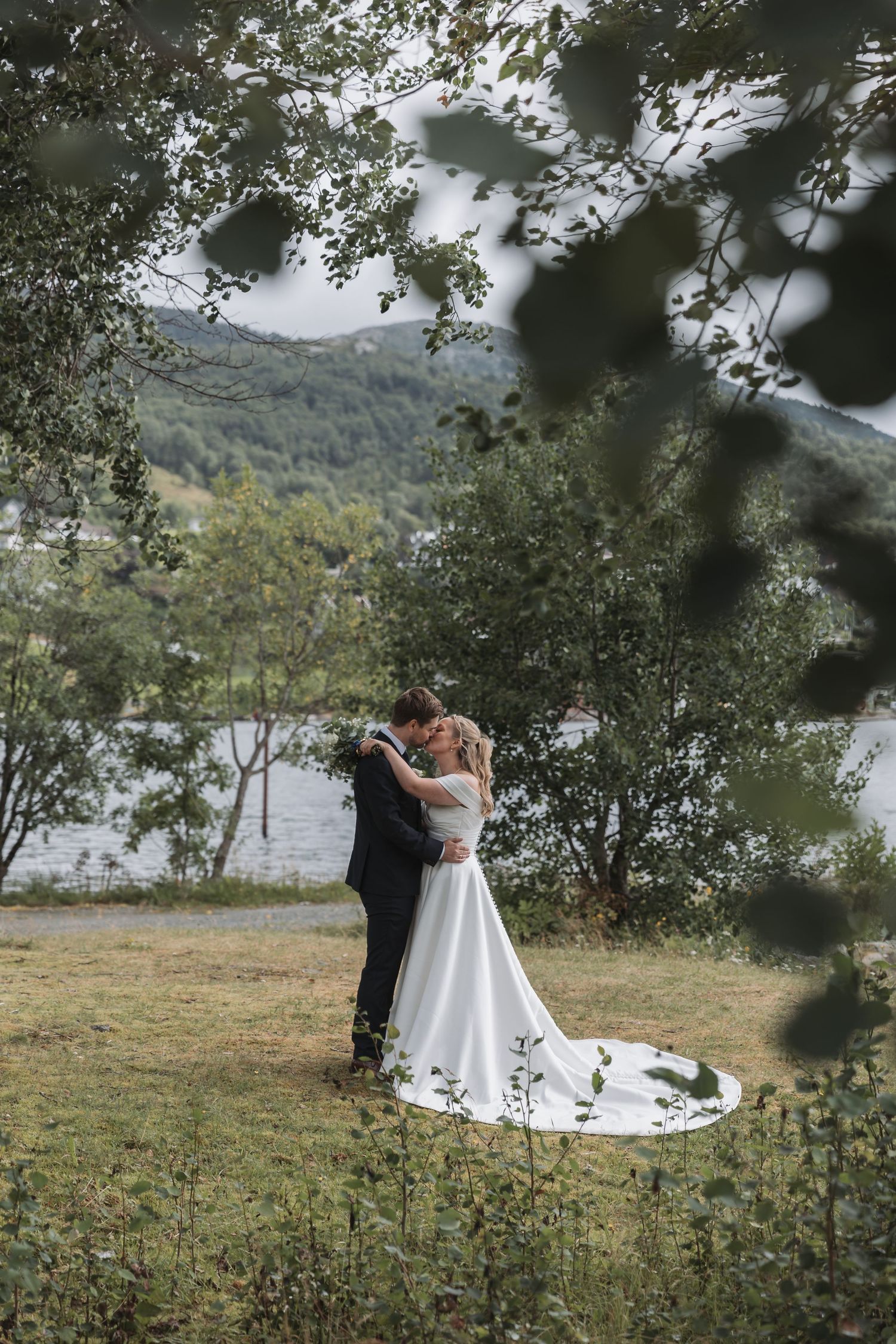 Bride and groom share a romantic kiss outdoors near a lake, surrounded by lush green trees on their wedding day.