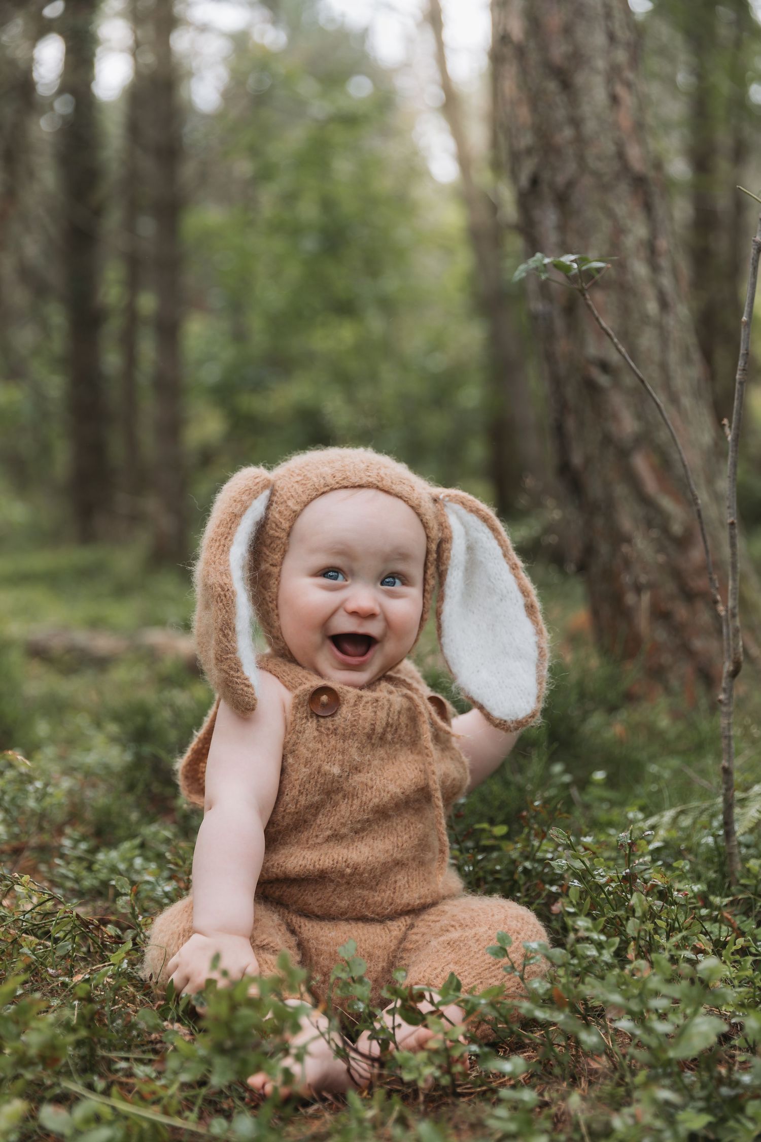 Small child wearing adorable brown bunny costume with floppy ears sits laughing in a forest setting.