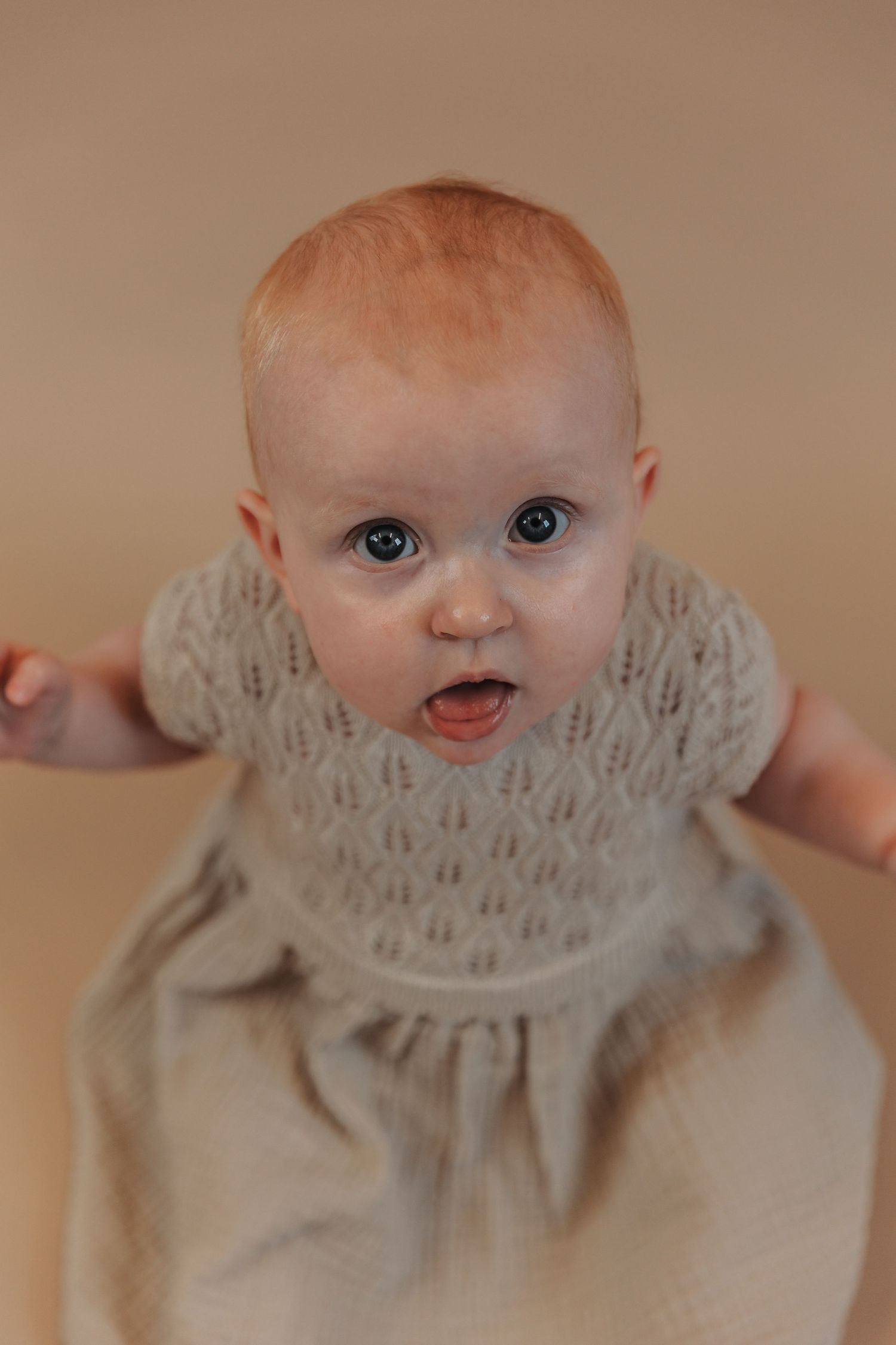 Baby wearing a beige dress with lace collar looks up at camera with bright blue eyes and surprised expression.