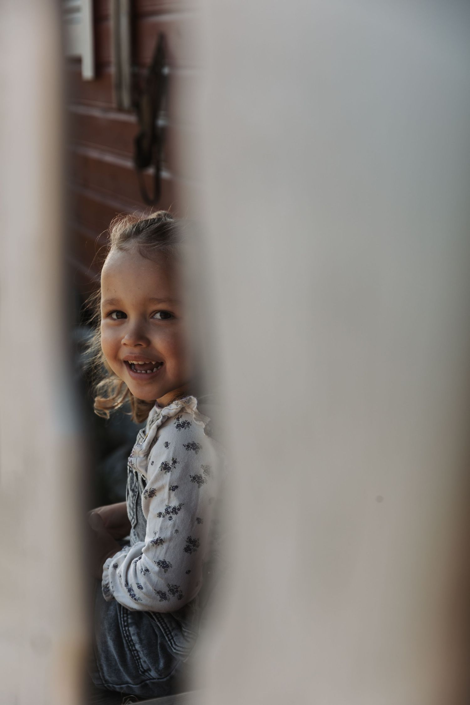 A view through a doorway shows a child with a bright smile wearing a patterned long sleeve shirt and dark skirt.
