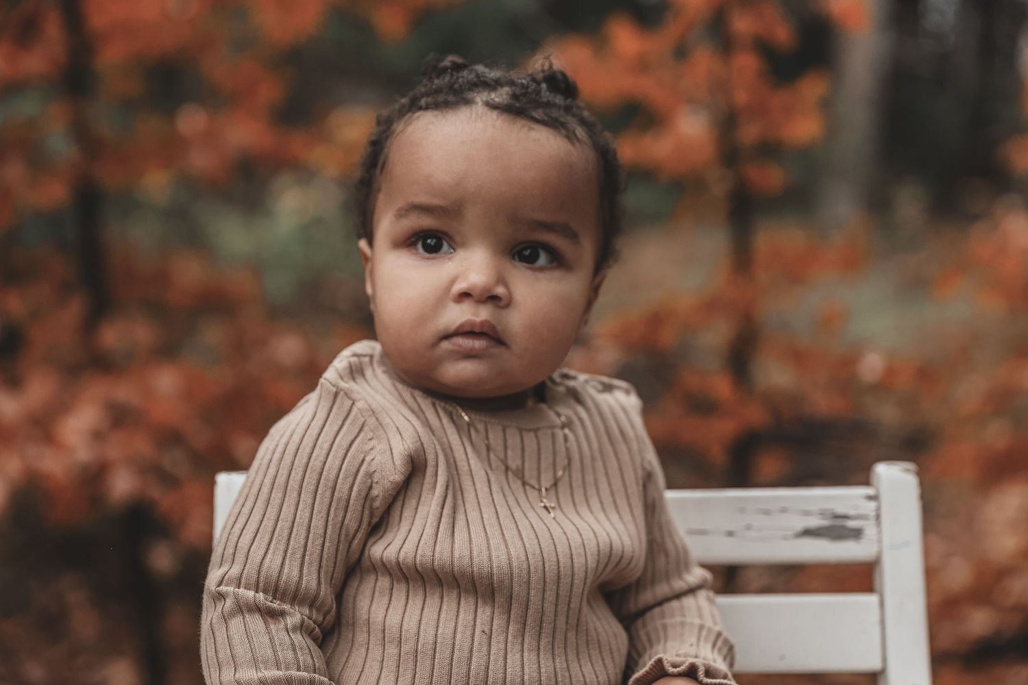 Small child wearing a brown sweater sits on white chair against autumn foliage background.