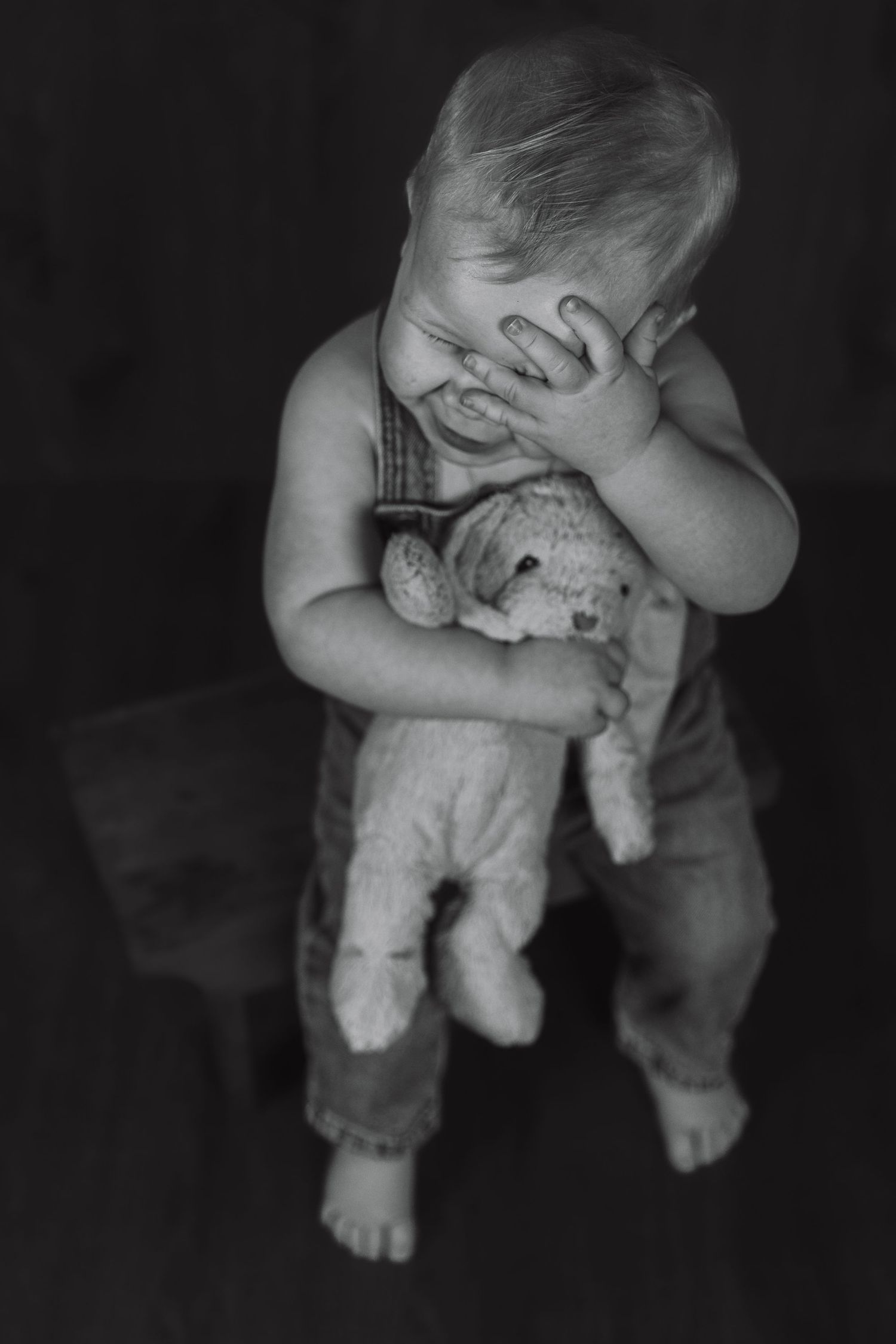 Black and white photo of toddler in overalls holding stuffed animal while covering face.