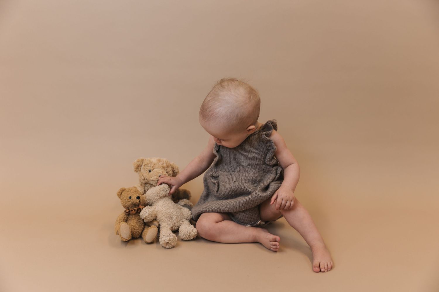 Baby in gray romper sitting on beige backdrop with teddy bear companion during studio photography session.
