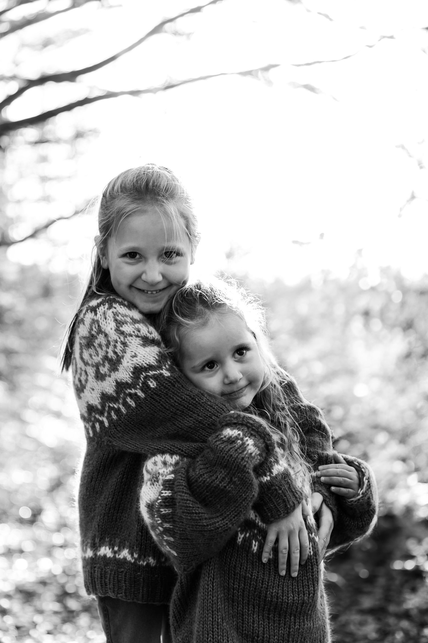 Black and white portrait of two children wearing cozy winter sweaters hugging and smiling outdoors in nature.