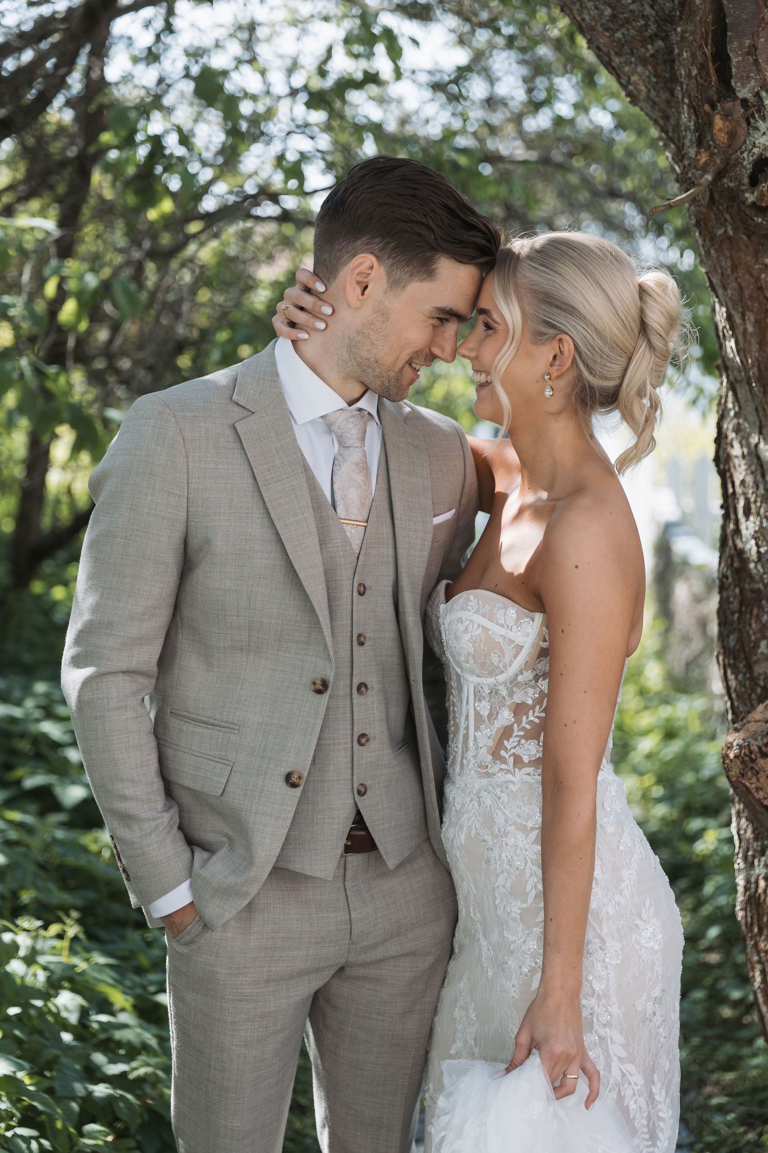 Bride and groom in elegant wedding attire share a romantic forehead touch surrounded by lush greenery outdoors.