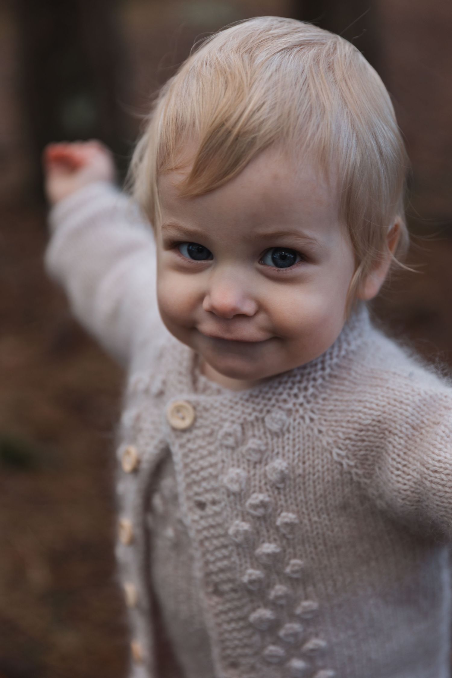Small child wearing a cozy knit sweater smiles while raising one arm in a playful gesture.