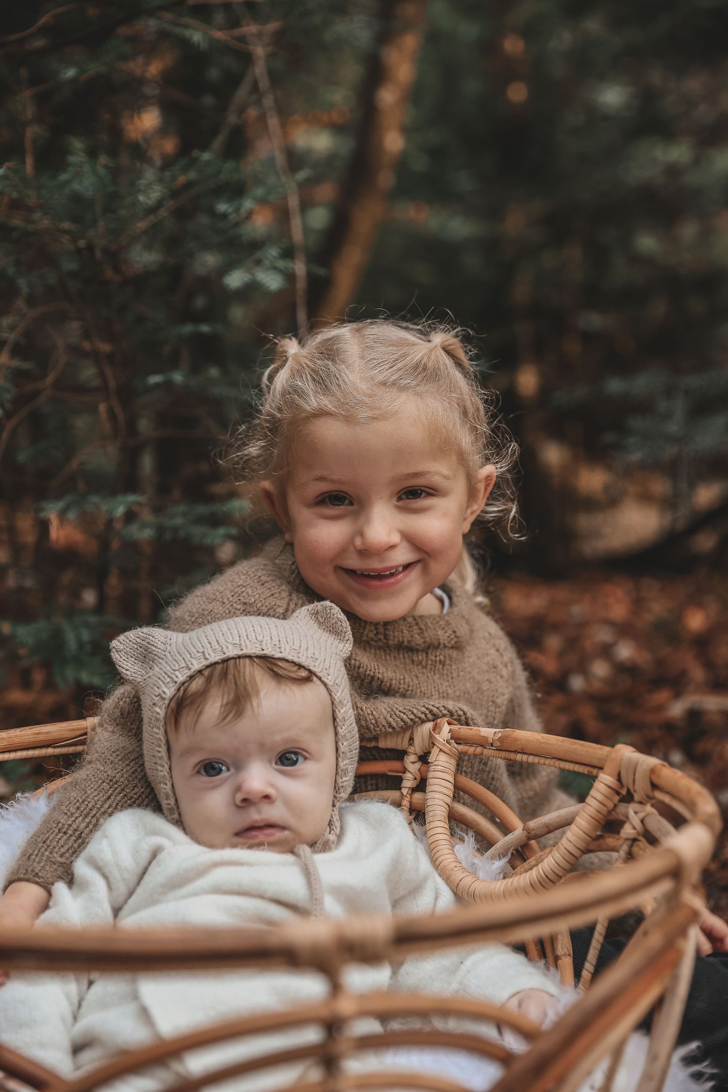 Two children in cozy knit sweaters sitting outdoors in a wicker chair surrounded by autumn foliage.
