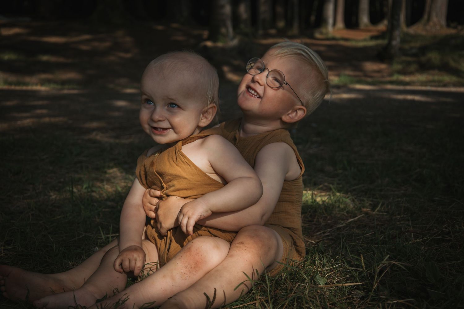 Two children embrace while sitting together in dappled forest light.