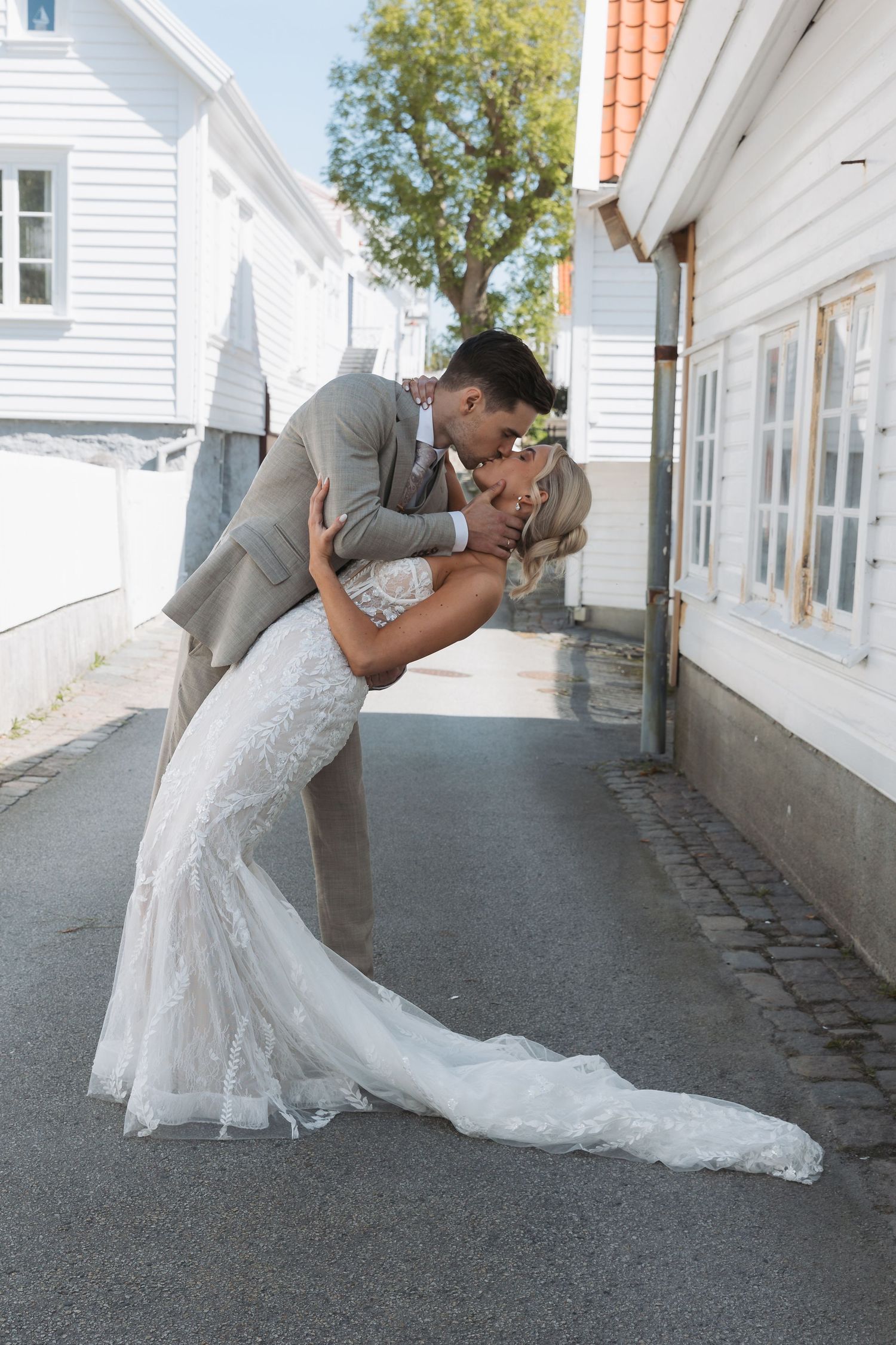Groom dips bride in lace mermaid wedding dress for romantic kiss on cobblestone street between white buildings.
