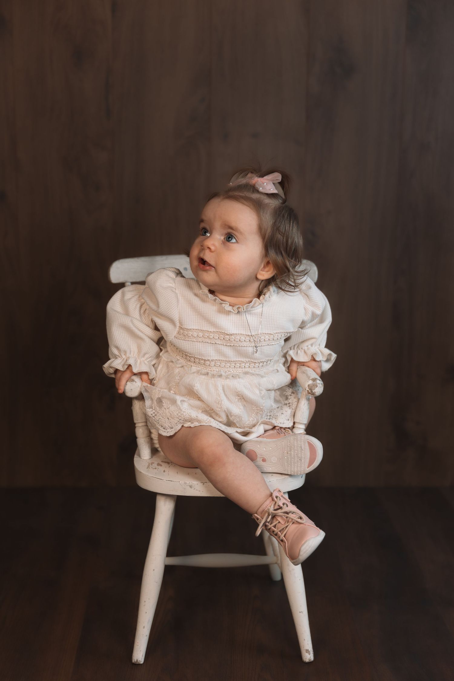 Small child in vintage-style ruffled dress sitting on white wooden chair against dark background.