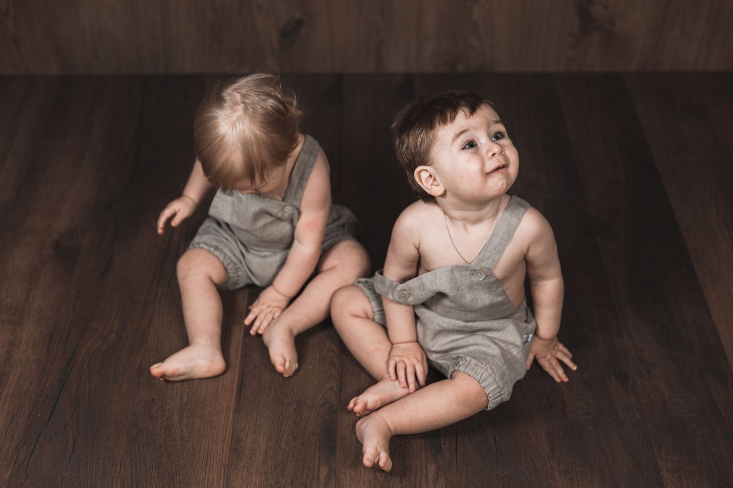 Two toddlers in matching gray rompers sitting on wooden flooring.