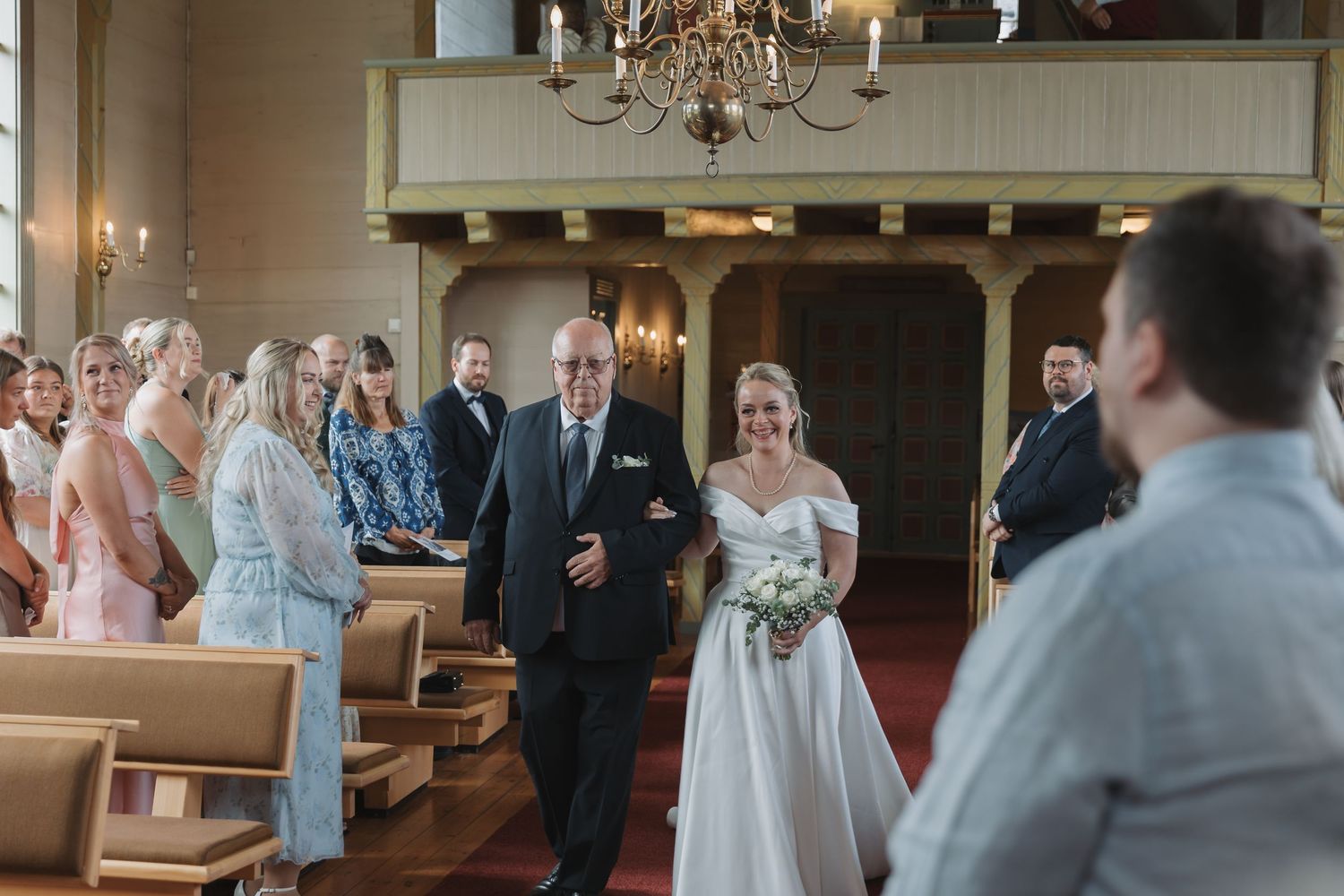 Bride walks down church aisle with escort, guests seated in wooden pews, chandelier overhead during wedding ceremony.
