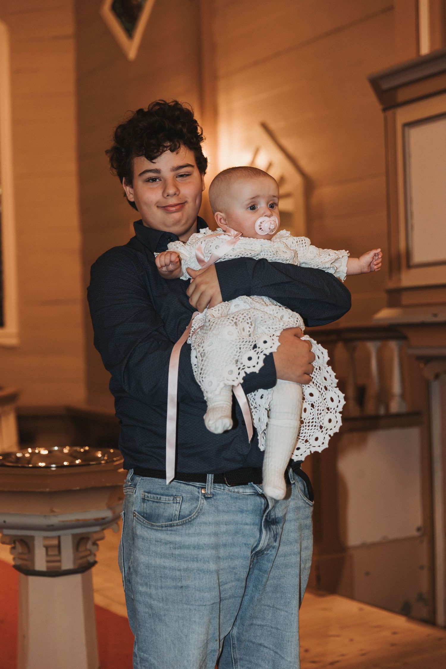 A baby wearing a white lacy dress is held by someone in a dark sweater and jeans in a warmly lit interior space.