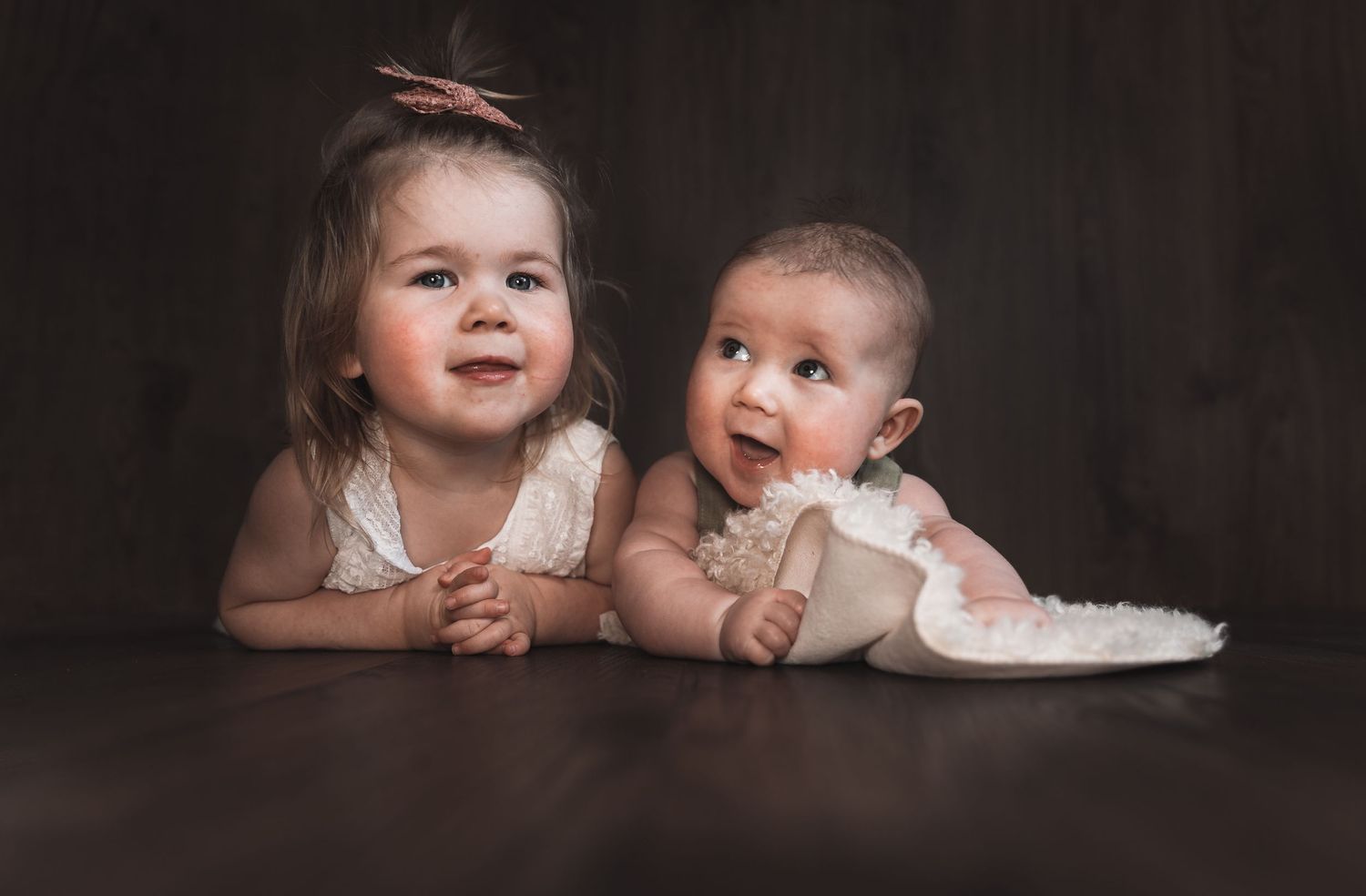 Two young children in white outfits lie on their stomachs against a dark brown backdrop for a studio portrait.