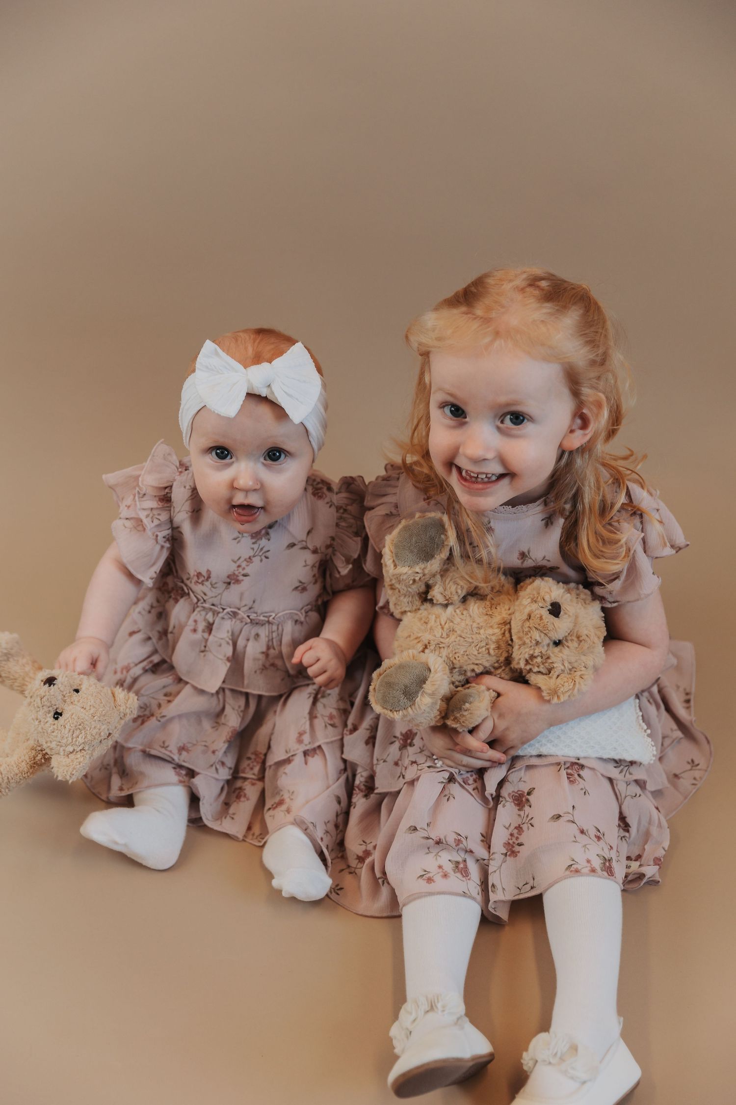 Two children in matching dusty rose dresses sitting together with teddy bears and white bows.